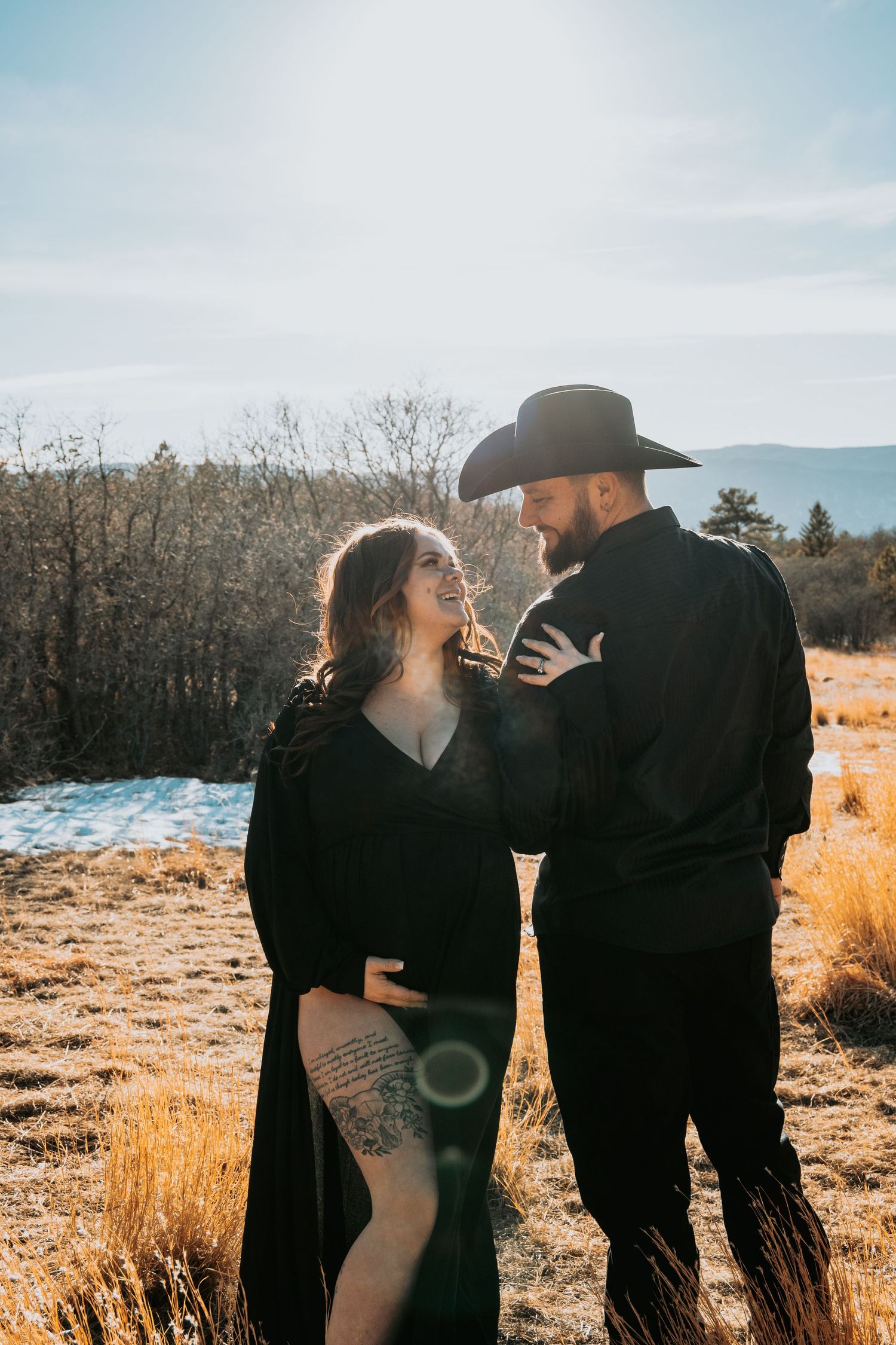 Smiling pregnant couple in black attire share a tender moment in sunlit winter field with mountains.