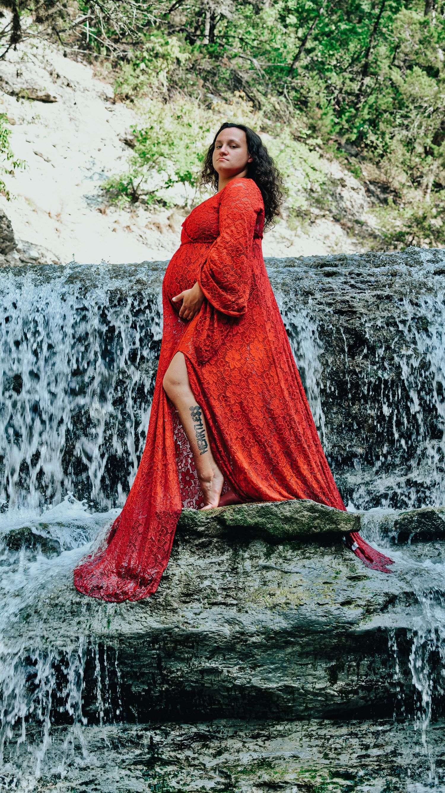 Pregnant woman in flowing red lace gown standing elegantly on rocks before a lush waterfall.