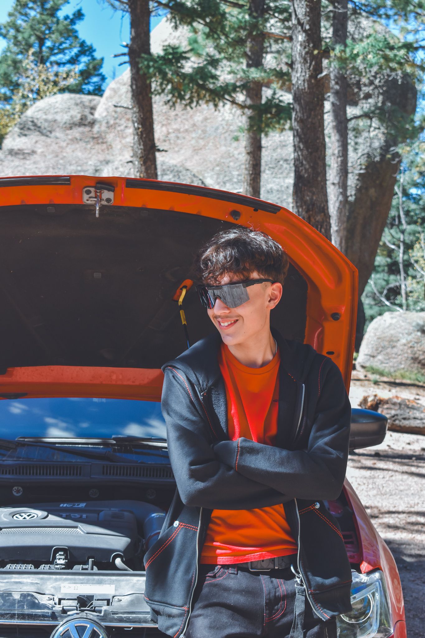 Smiling teen in red shirt and black jacket leaning on orange car with open hood in forest for senior photos.