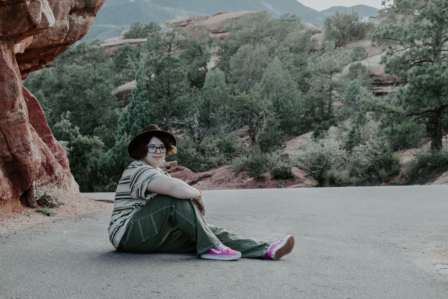 Person sitting on a road near red rock formations, wearing a striped outfit, hat, and pink shoes for senior photos.