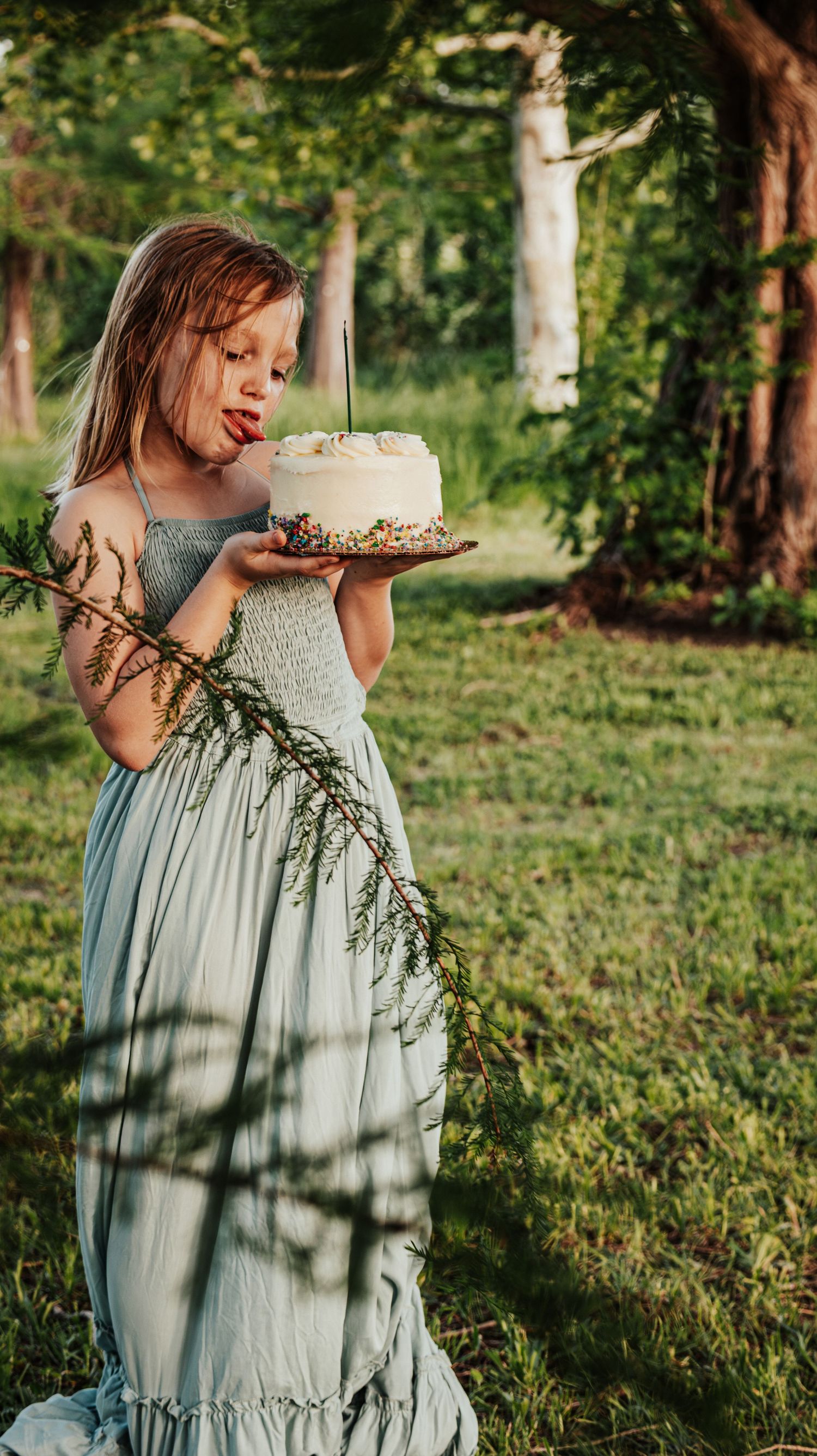 A girl in a long sage green dress blows a candle on a white birthday cake in a lush garden.