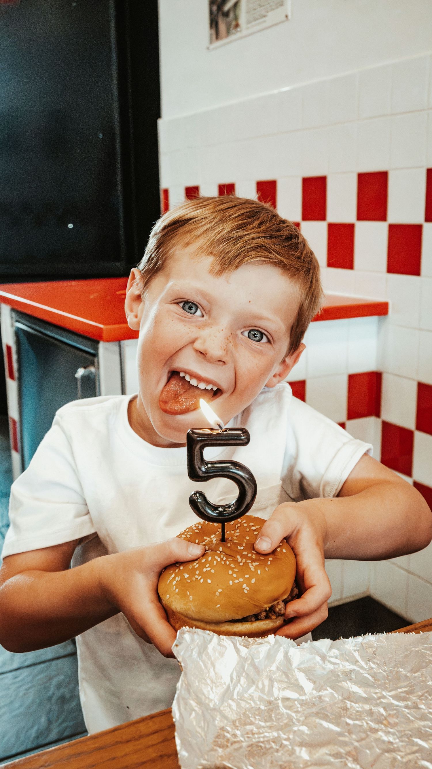 Excited young boy holding a burger with a number 5 birthday candle at Five Guys restaurant.