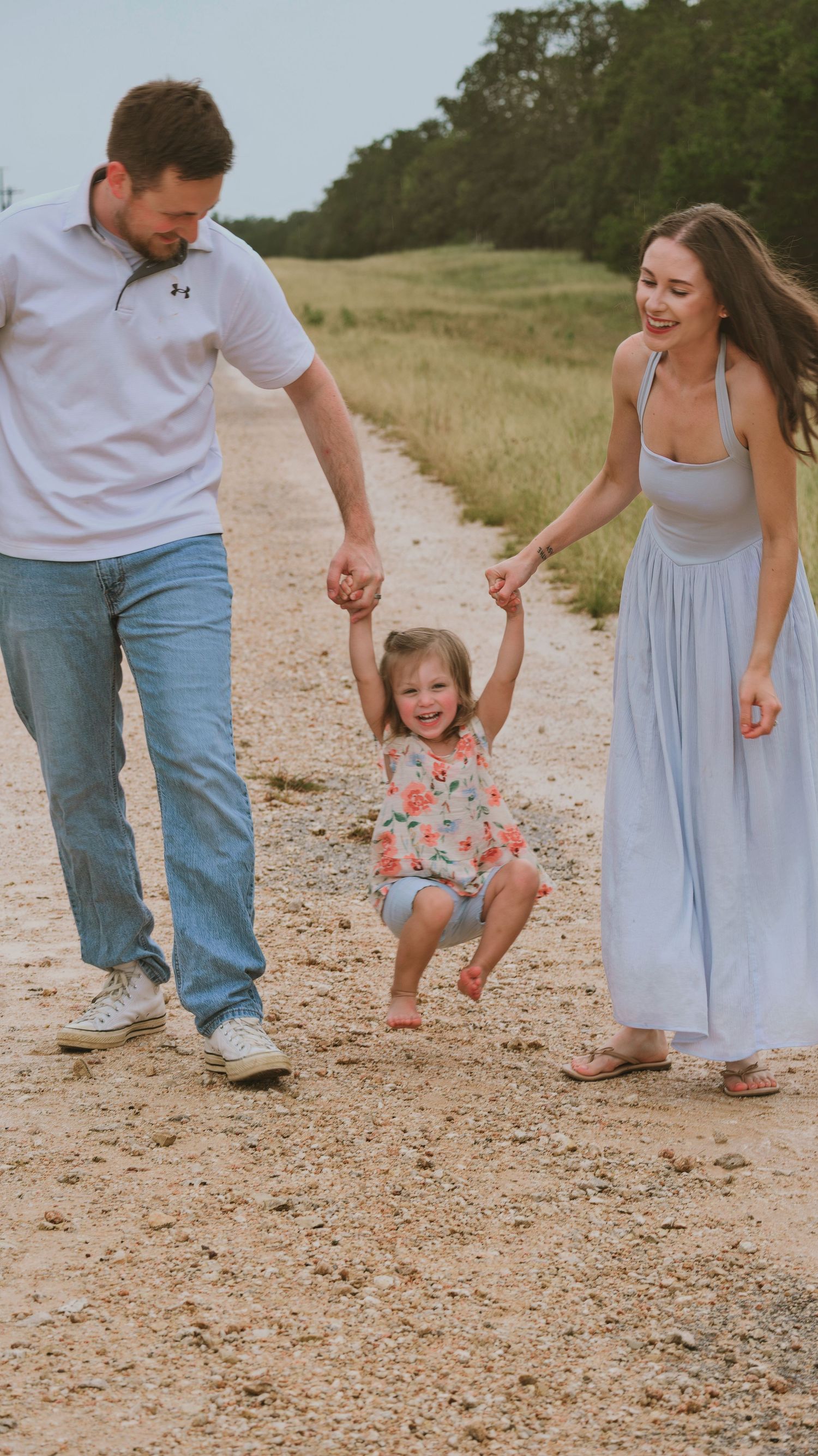Happy family swings their laughing toddler girl on a sunny dirt path for a family session.