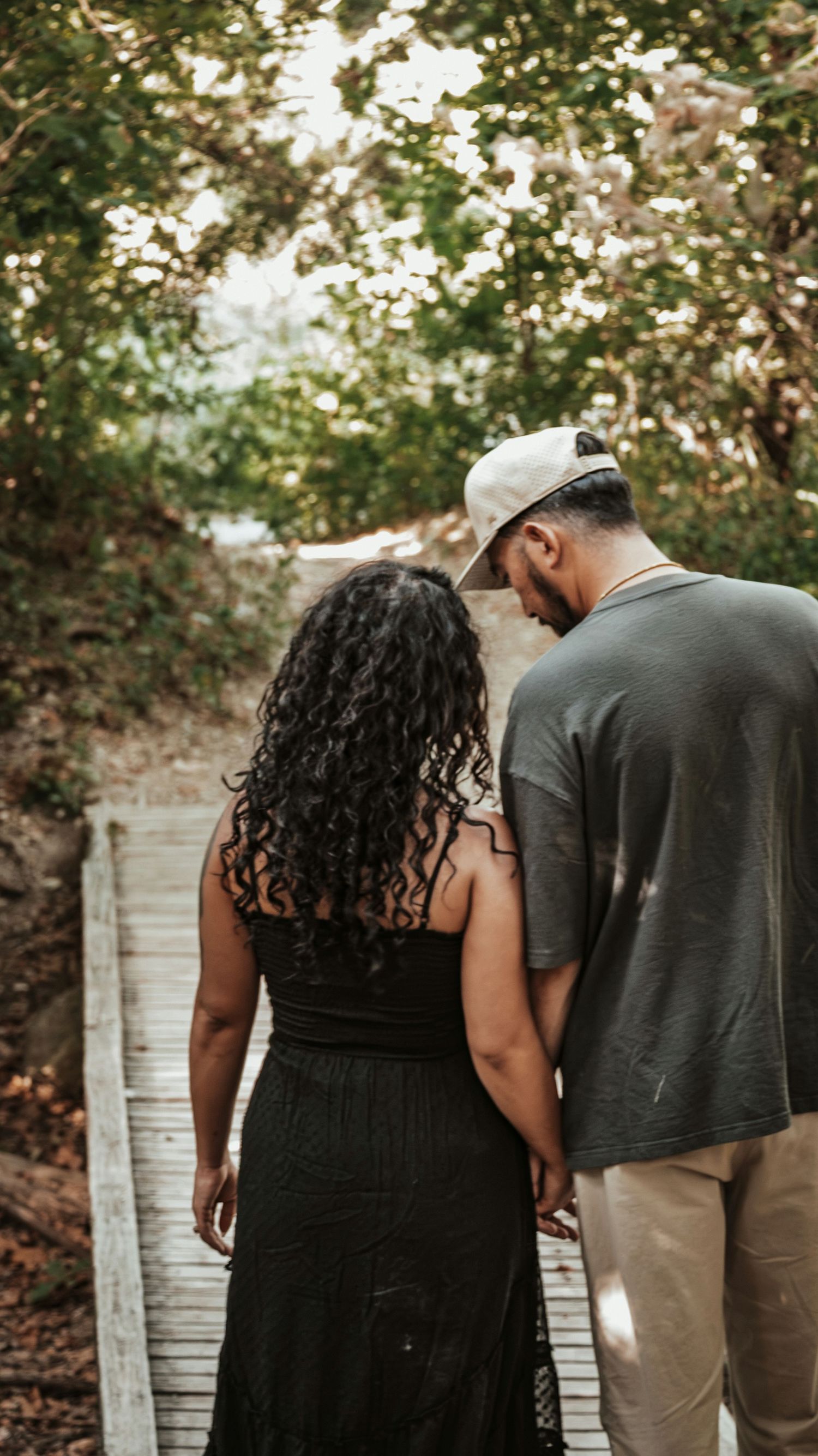 A couple walks hand-in-hand down a wooden path surrounded by lush green trees, viewed from behind for a couples session.