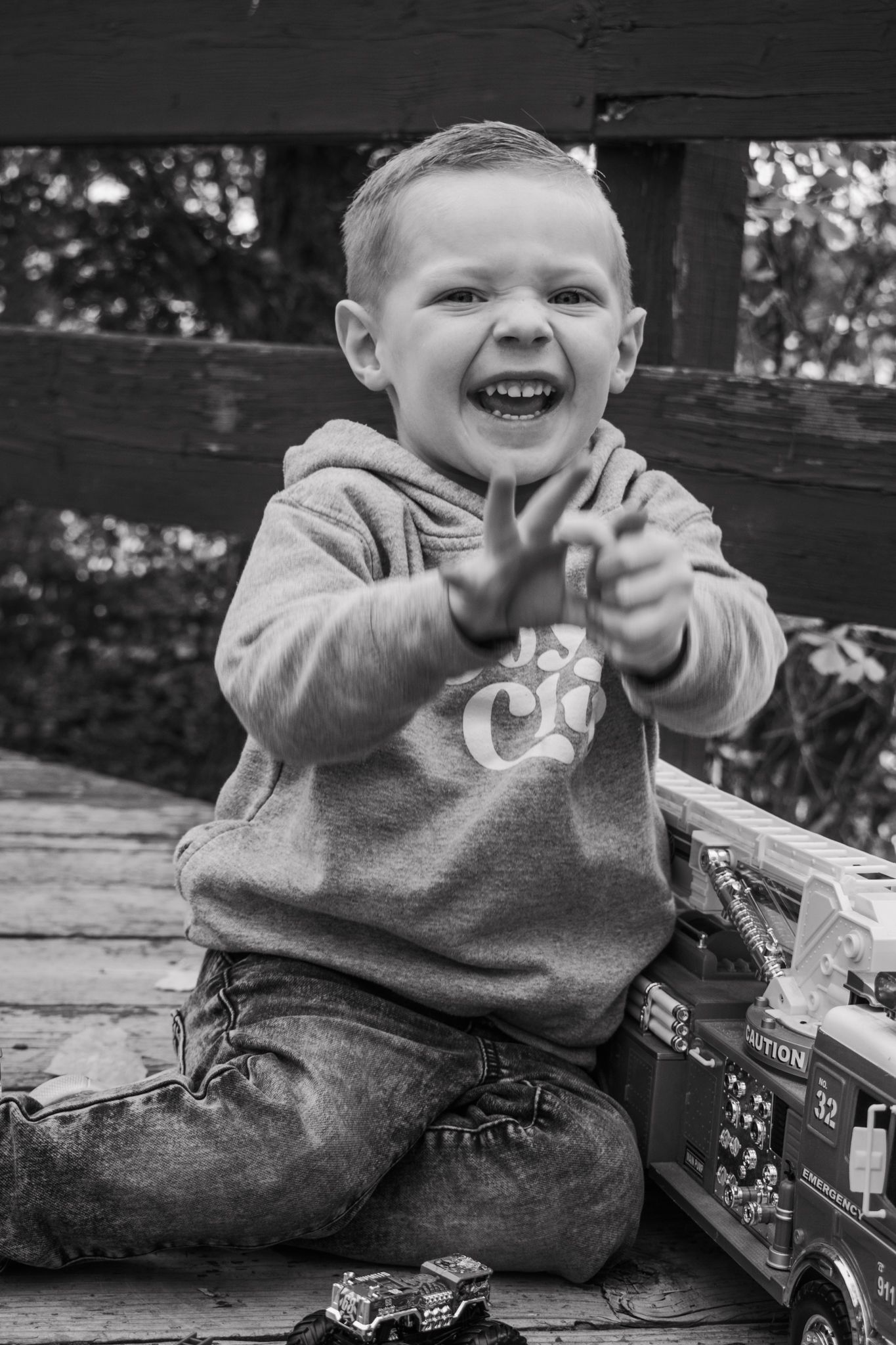 Happy laughing toddler boy playing with toy fire trucks on a wooden deck in black and white. Family Photographer in Pflugerville, Tx