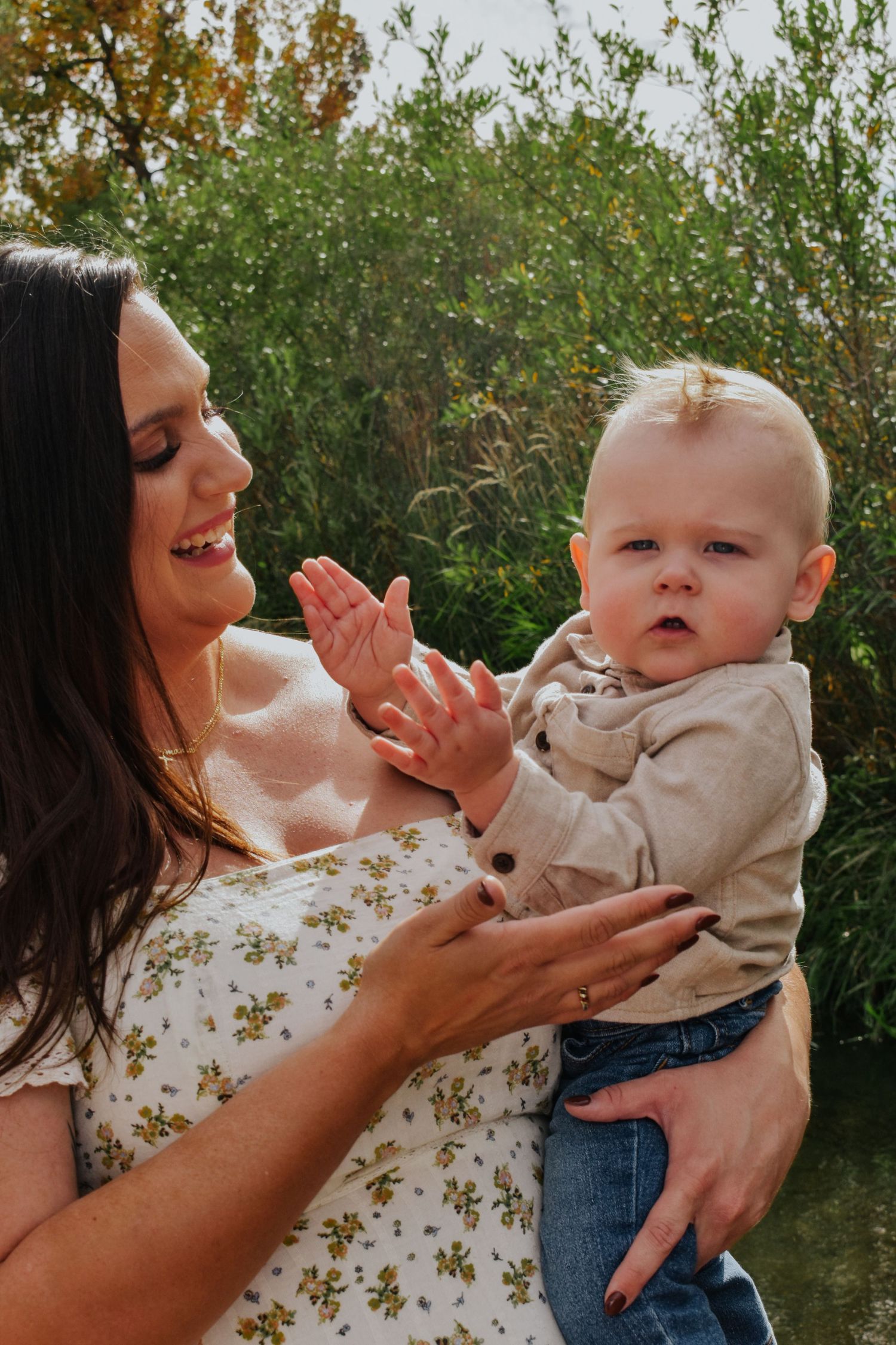 Smiling brunette mother in floral dress holding her baby boy outdoors.
