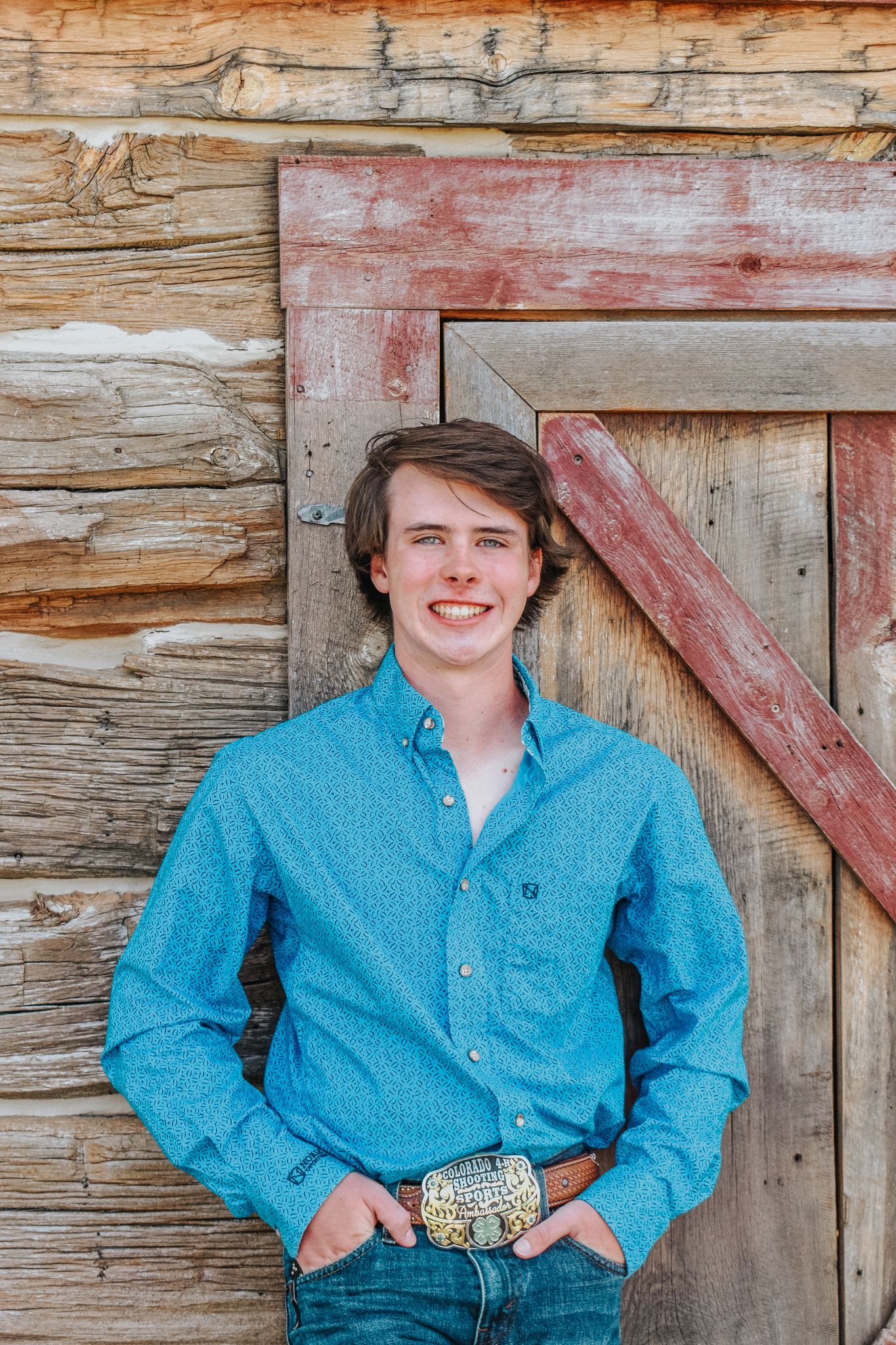 Smiling young man in teal western shirt with decorative belt buckle leaning against rustic wooden barn door for senior photos.