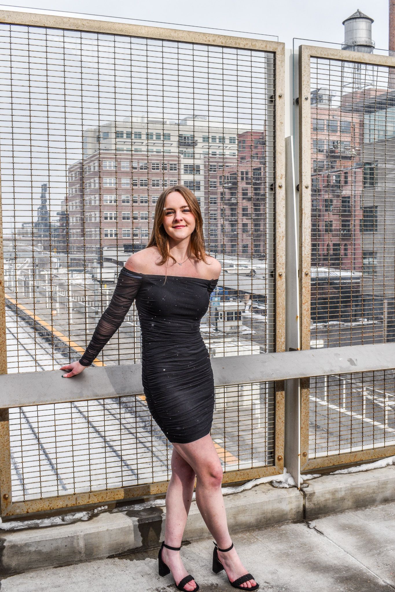 Young woman in black off-shoulder dress leaning on rooftop fence with urban cityscape behind her for senior photos.