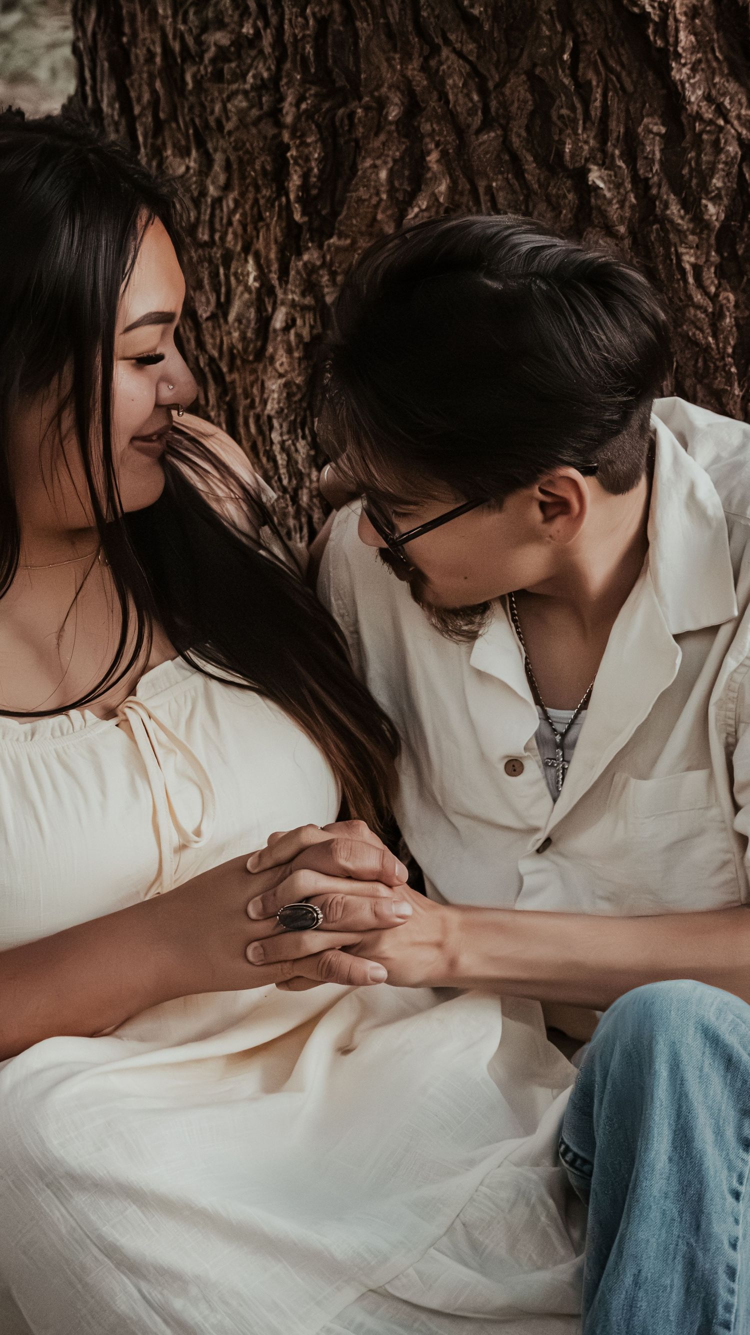 A couple sits together against a tree as the man gently kisses the womans cheek.