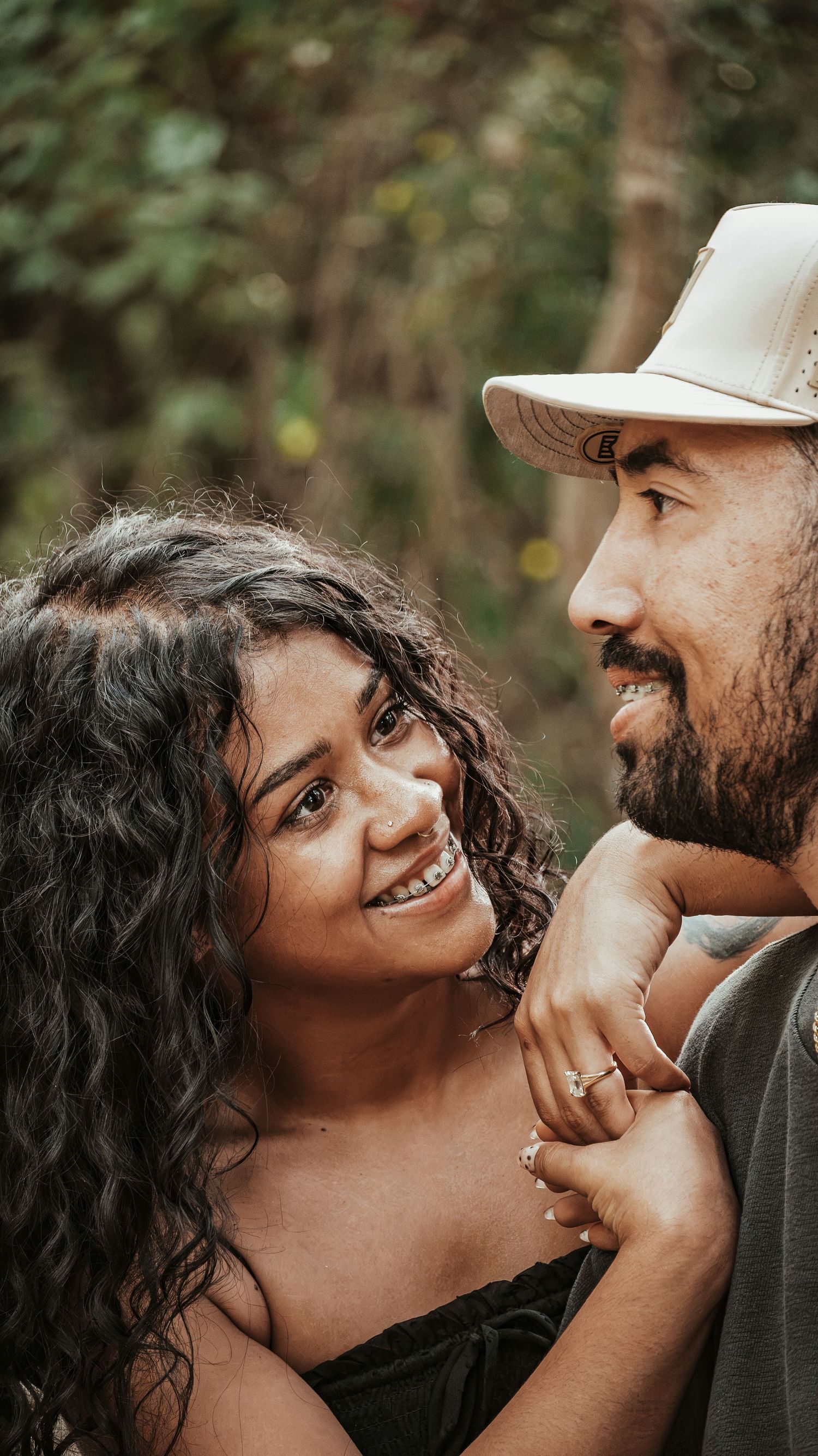 A happy couple smiles at each other outdoors, the woman wearing a ring in a forest.