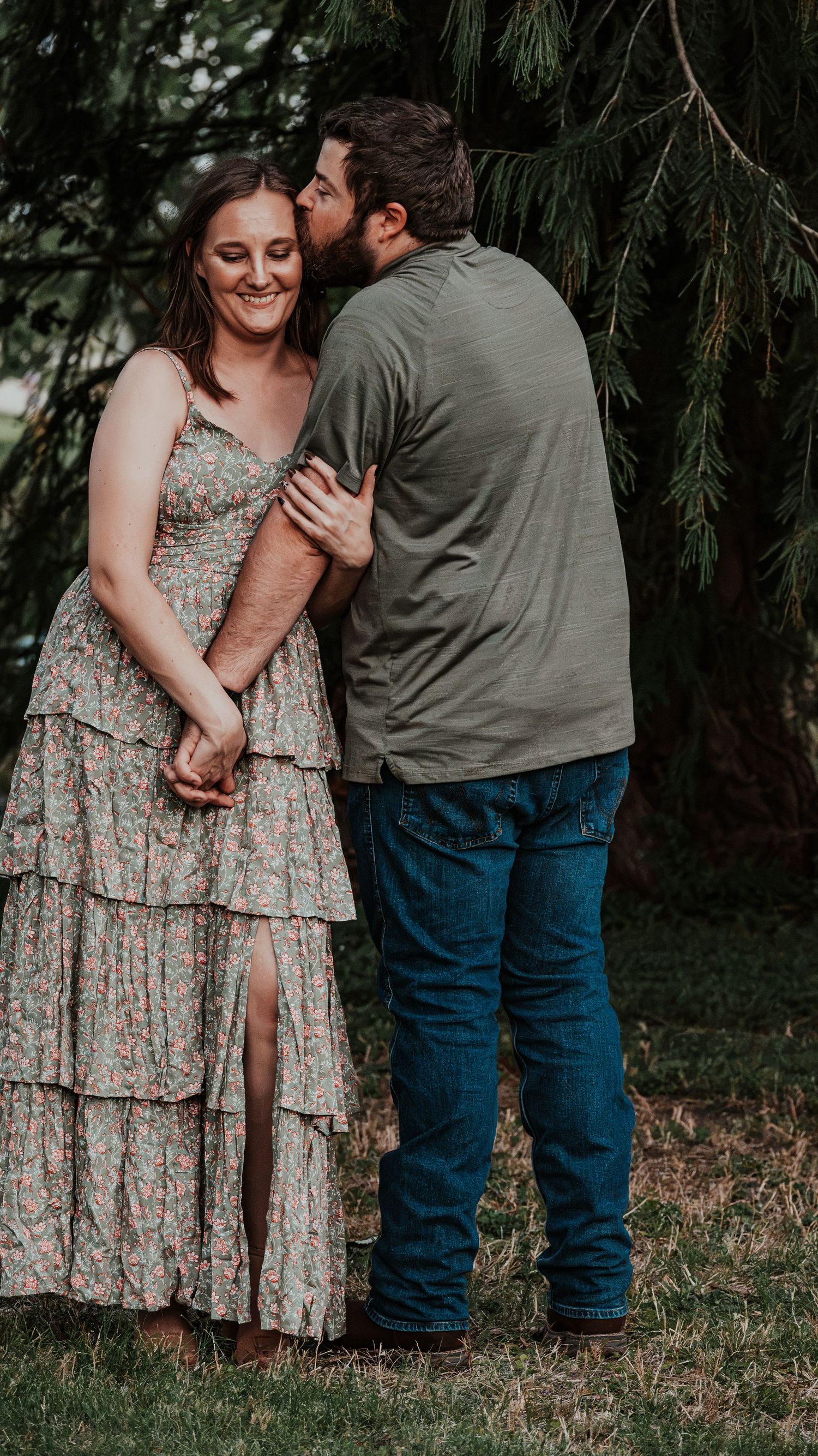 A couple stands closely together under a pine tree as the man kisses the smiling woman on the cheek.