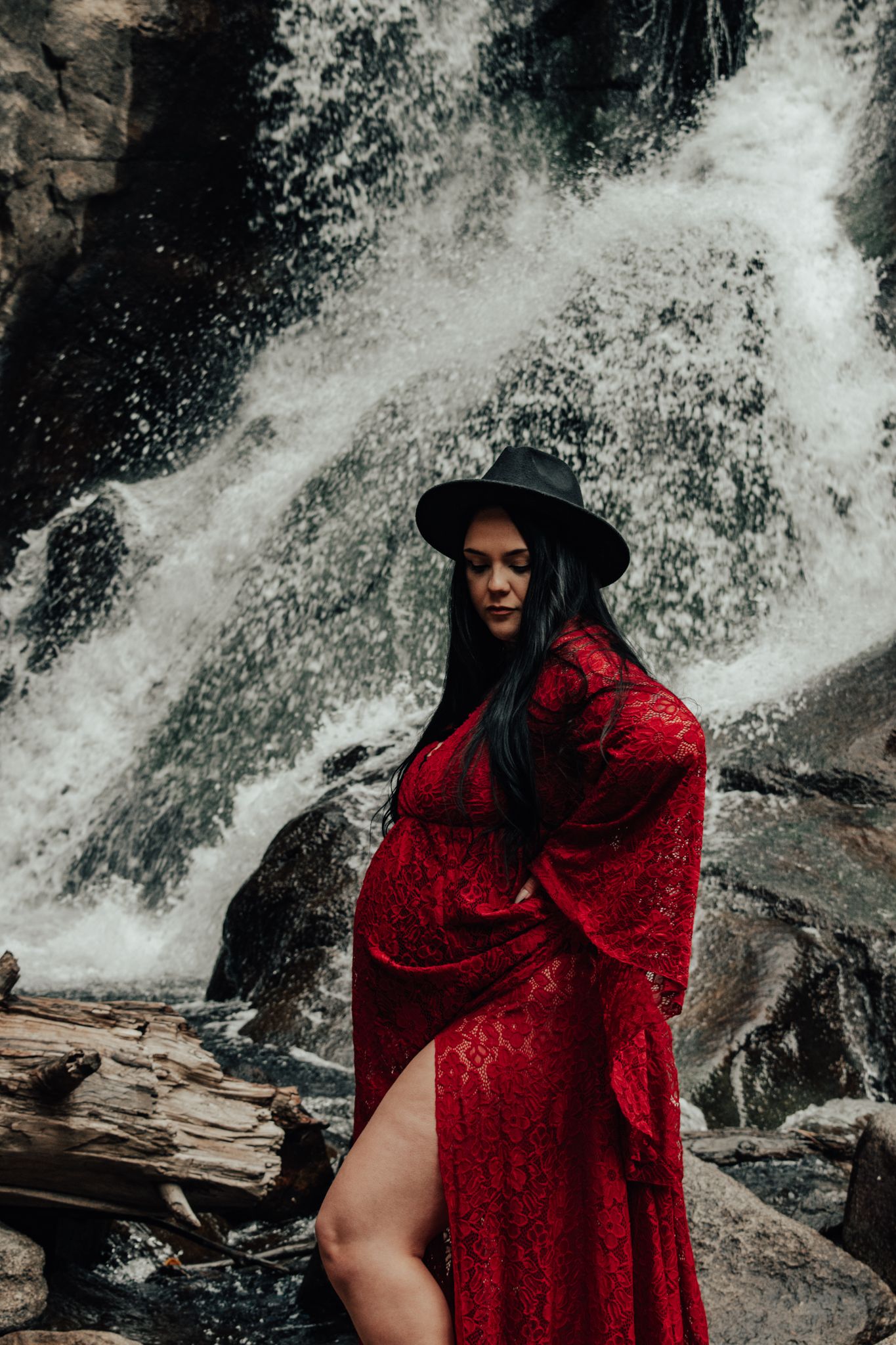 Pregnant woman in red lace dress and wide-brim hat stands pensively before cascading waterfall.