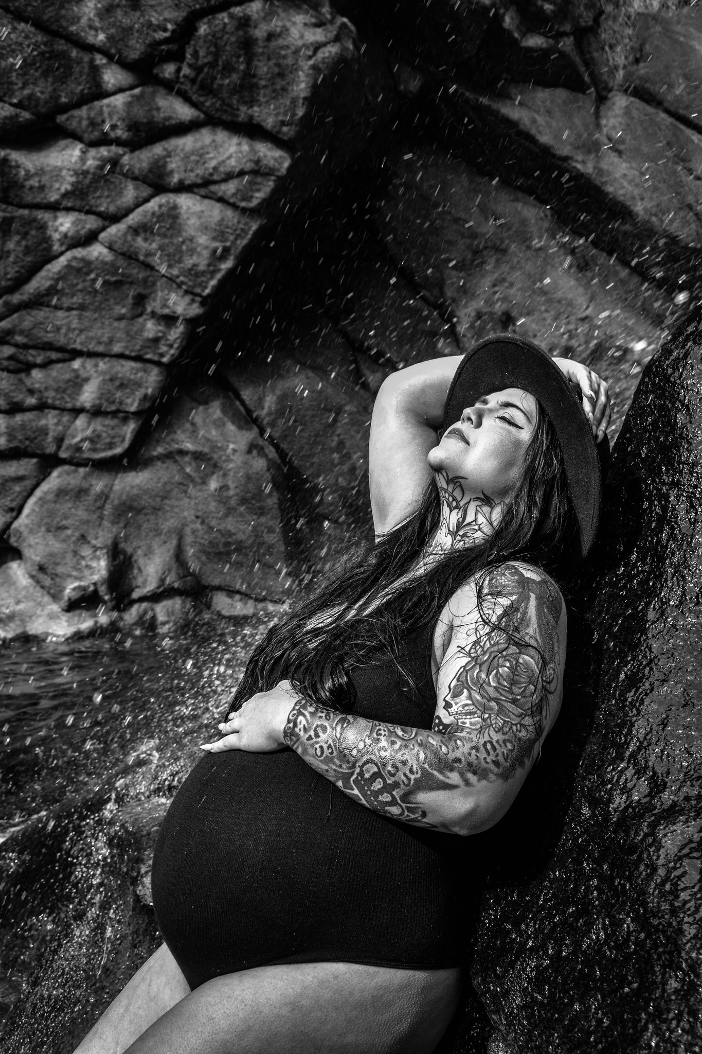 Pregnant tattooed woman in black swimsuit leaning against rocks near waterfall. Maternity Photographer in Pflugerville, Tx