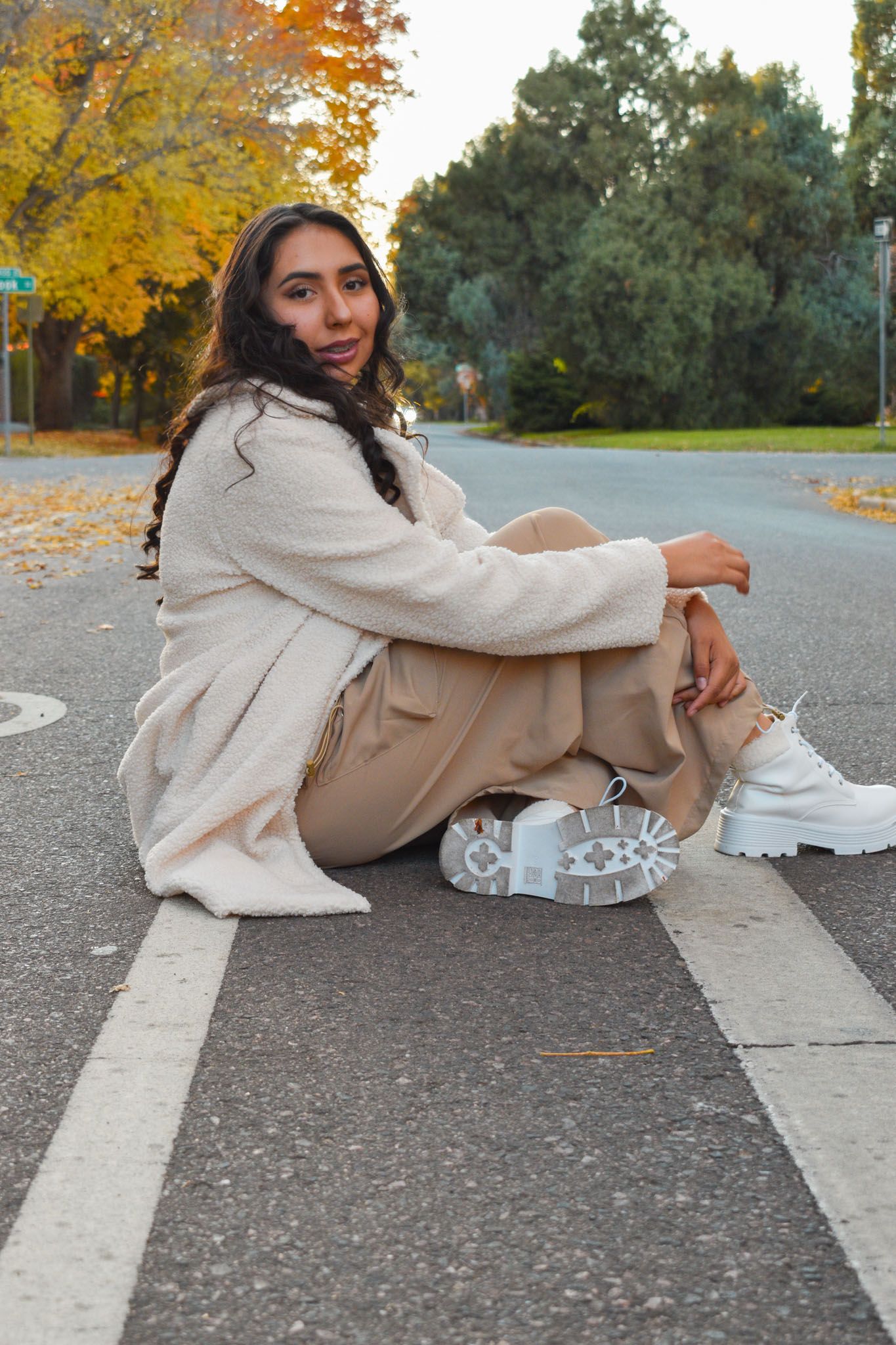 Young woman in beige coat and tan pants sitting on road stripe during autumn sunset for senior photos.