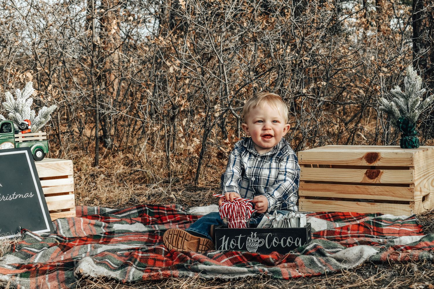 A smiling toddler boy in a plaid shirt sits on a red tartan blanket with Christmas decorations outdoors.
