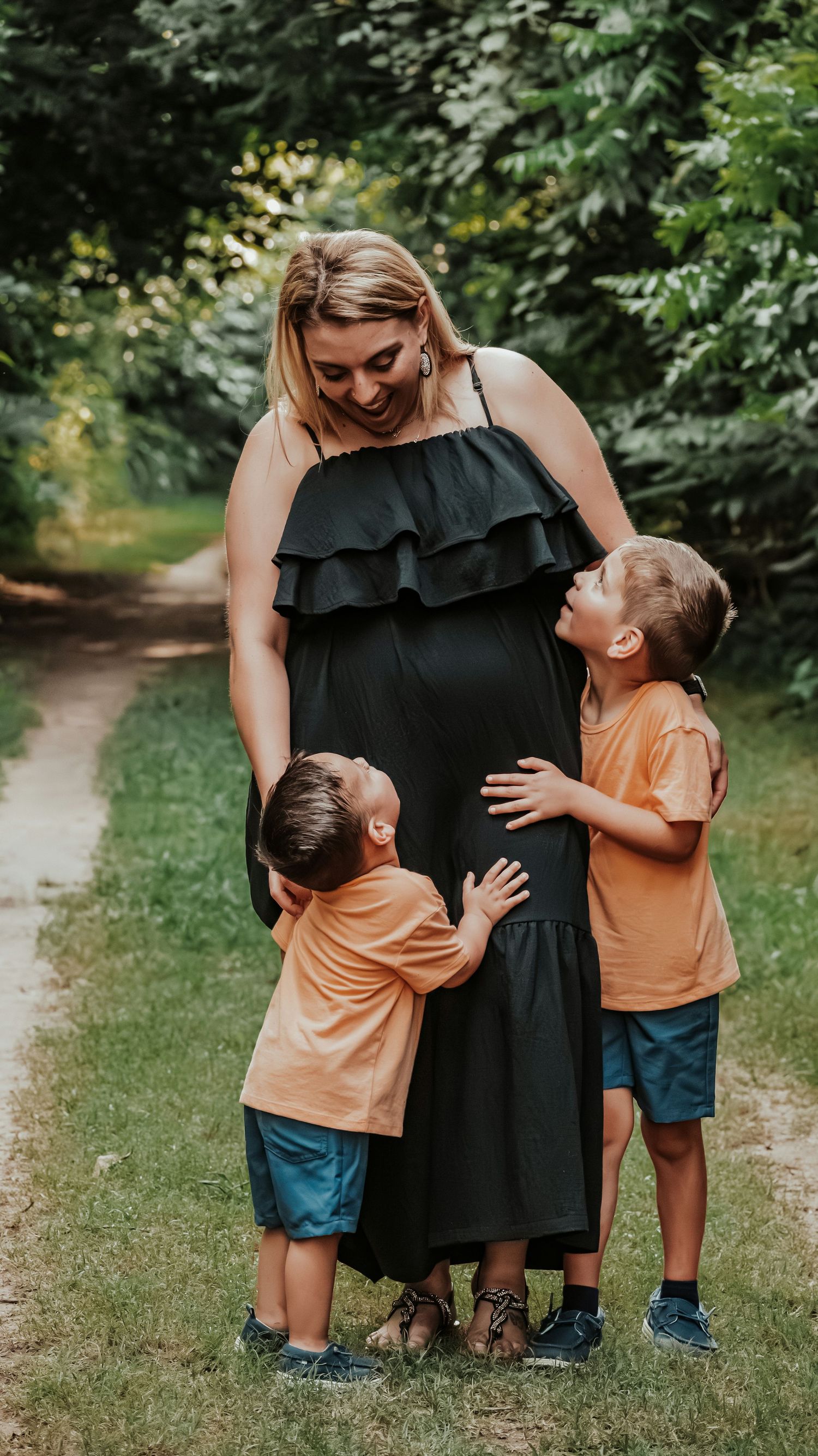 Two young boys in orange shirts hugging their pregnant mothers baby bump on a scenic green nature path.