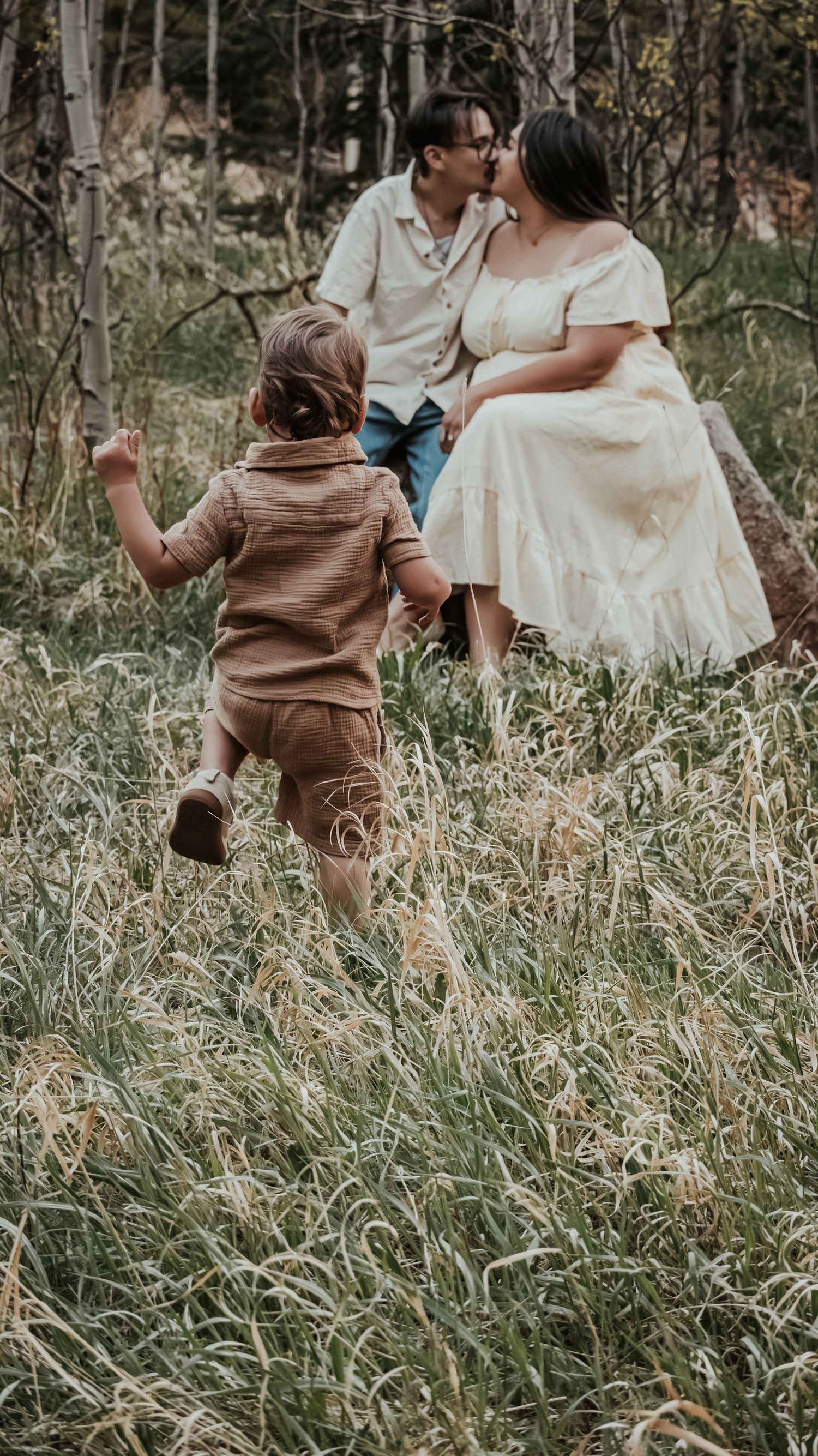 A toddler runs through tall grass toward his parents who share a kiss in a forest meadow.
