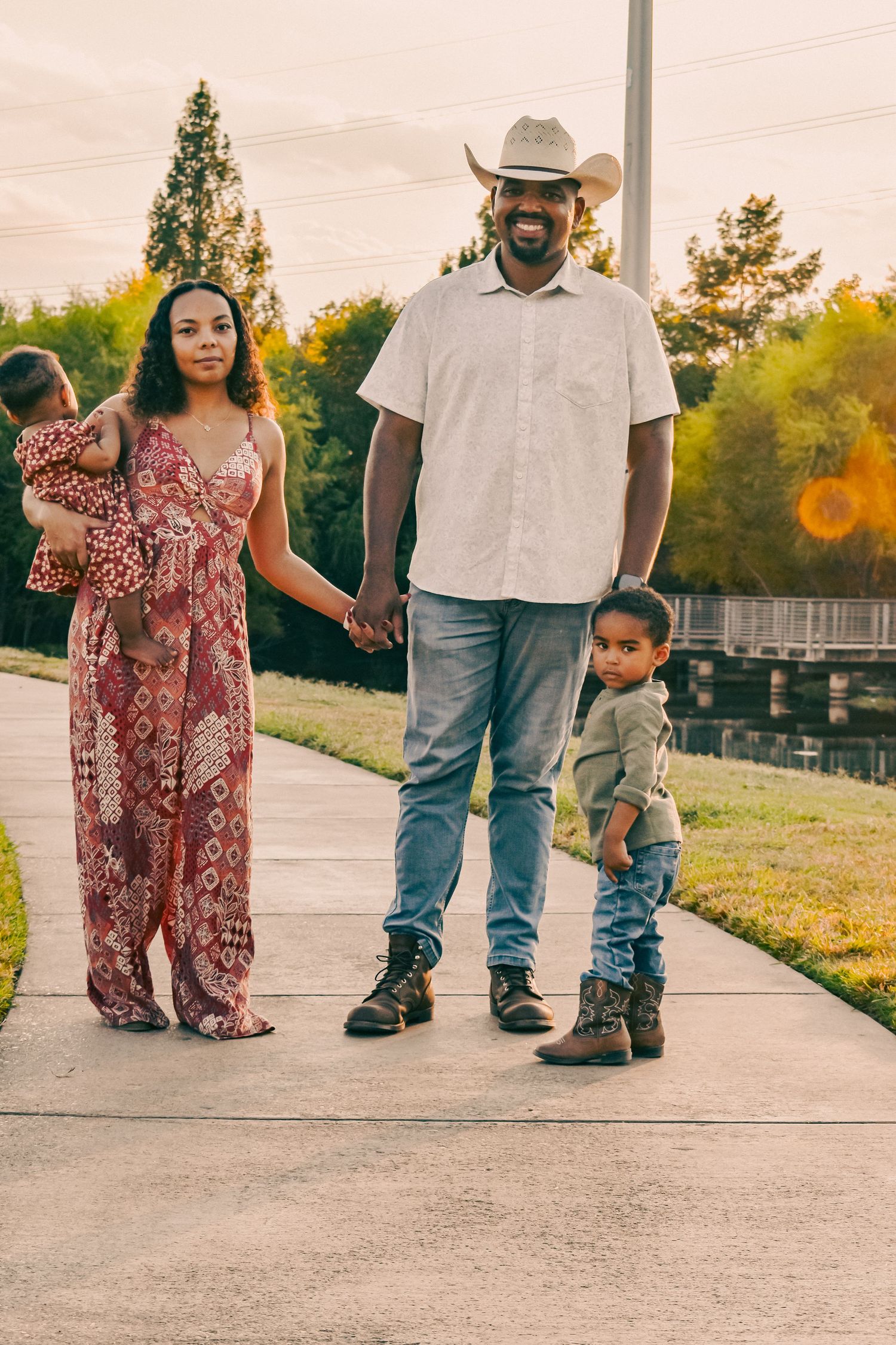 Smiling Black family of four poses on a park sidewalk at sunset, dad in cowboy hat, mom in floral jumpsuit with two kids.