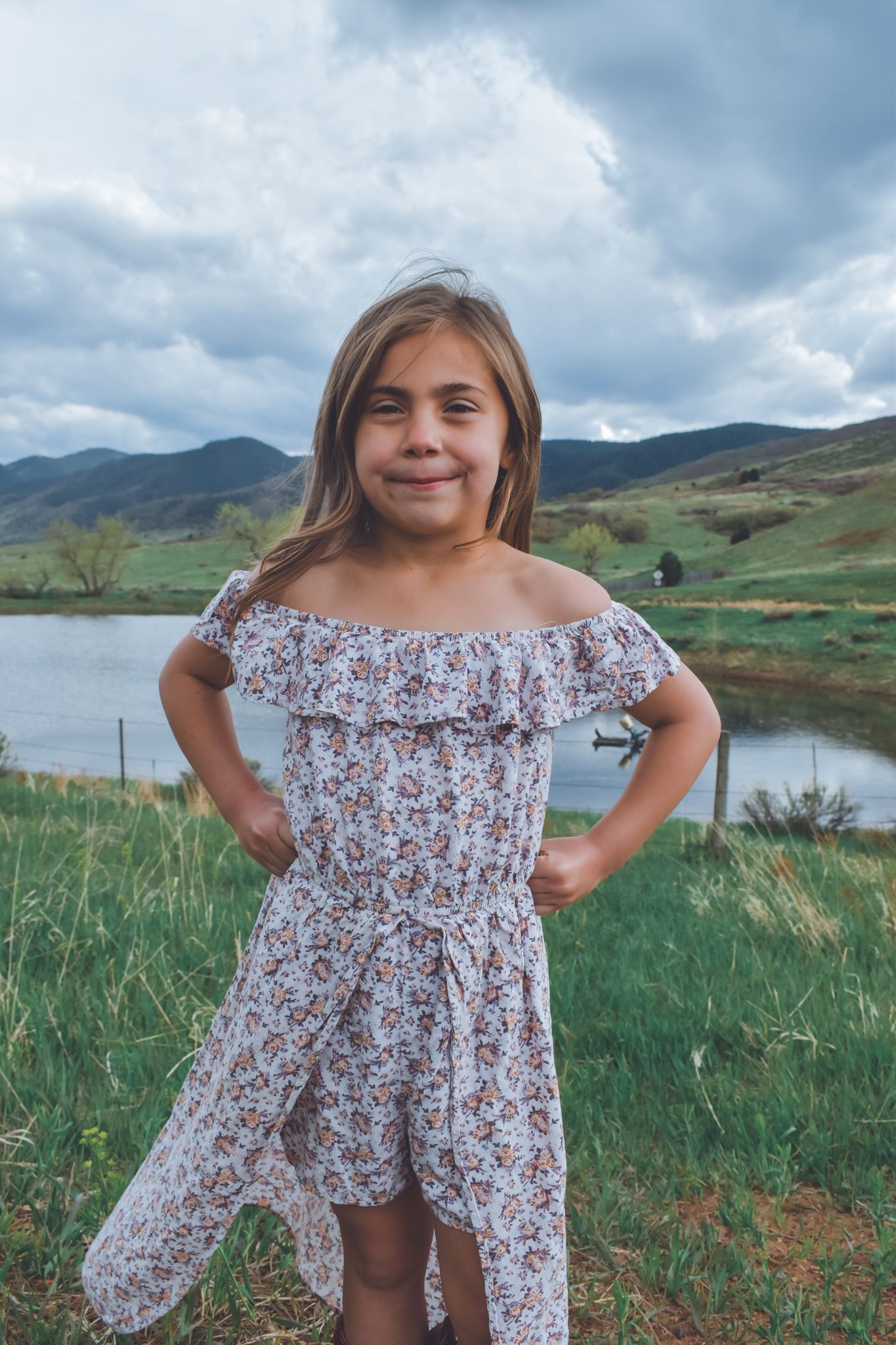 Happy girl in floral off-shoulder dress posing with hands on hips near mountain lake.