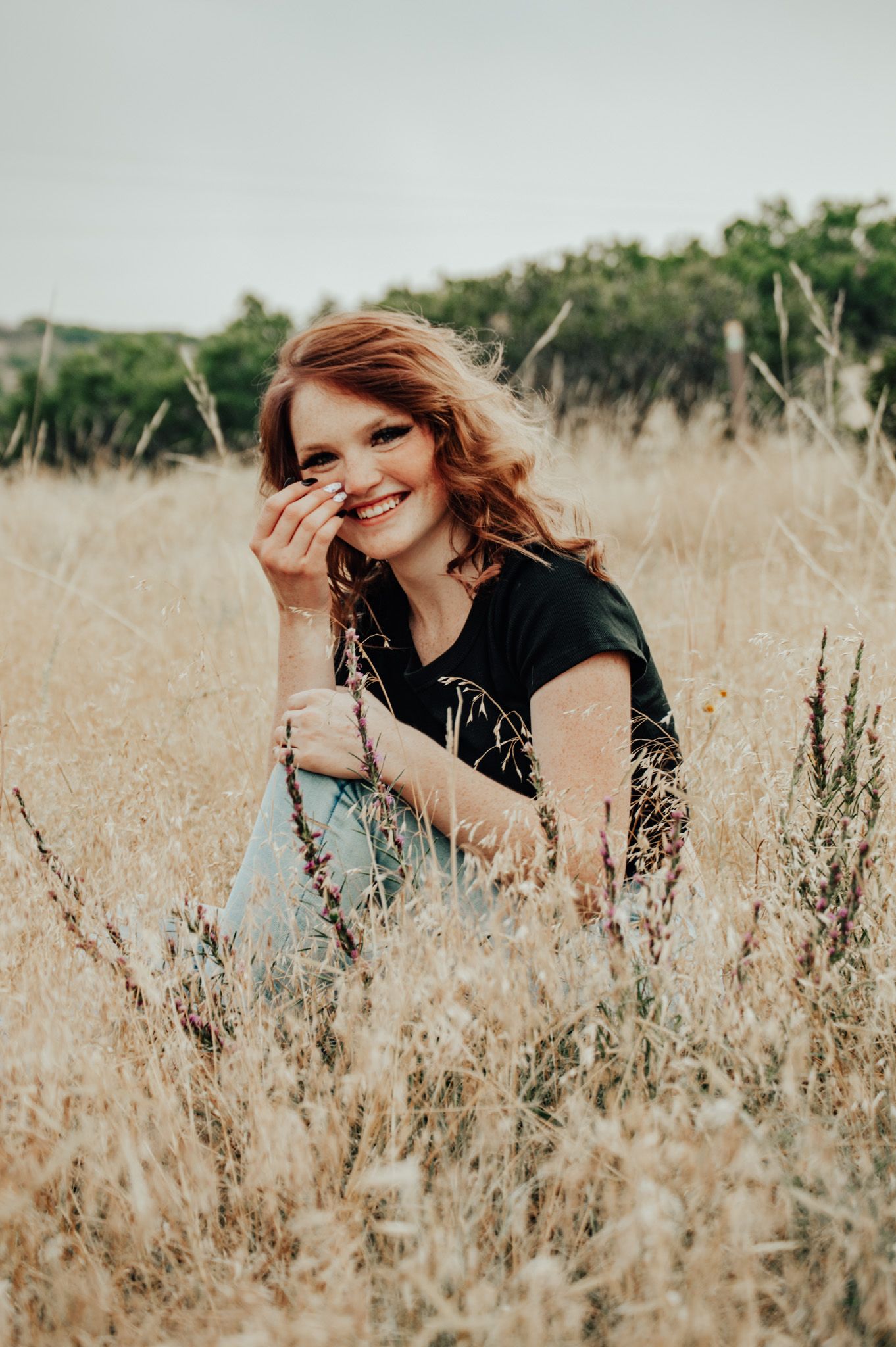 Laughing redhead woman sitting in golden field grass wearing black t-shirt and light blue jeans for senior photos.