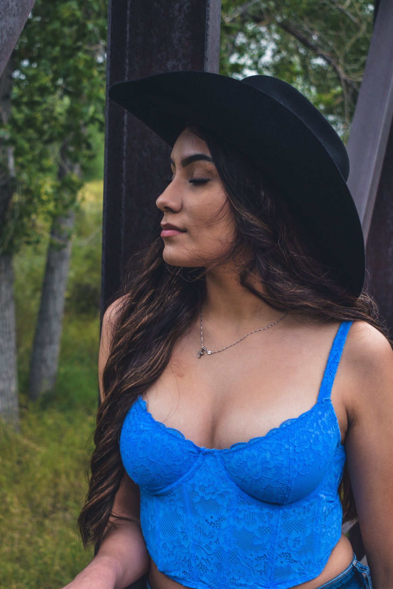 Young woman in blue lace crop top and black wide-brim hat poses near rustic metal structure outdoors for senior photos.