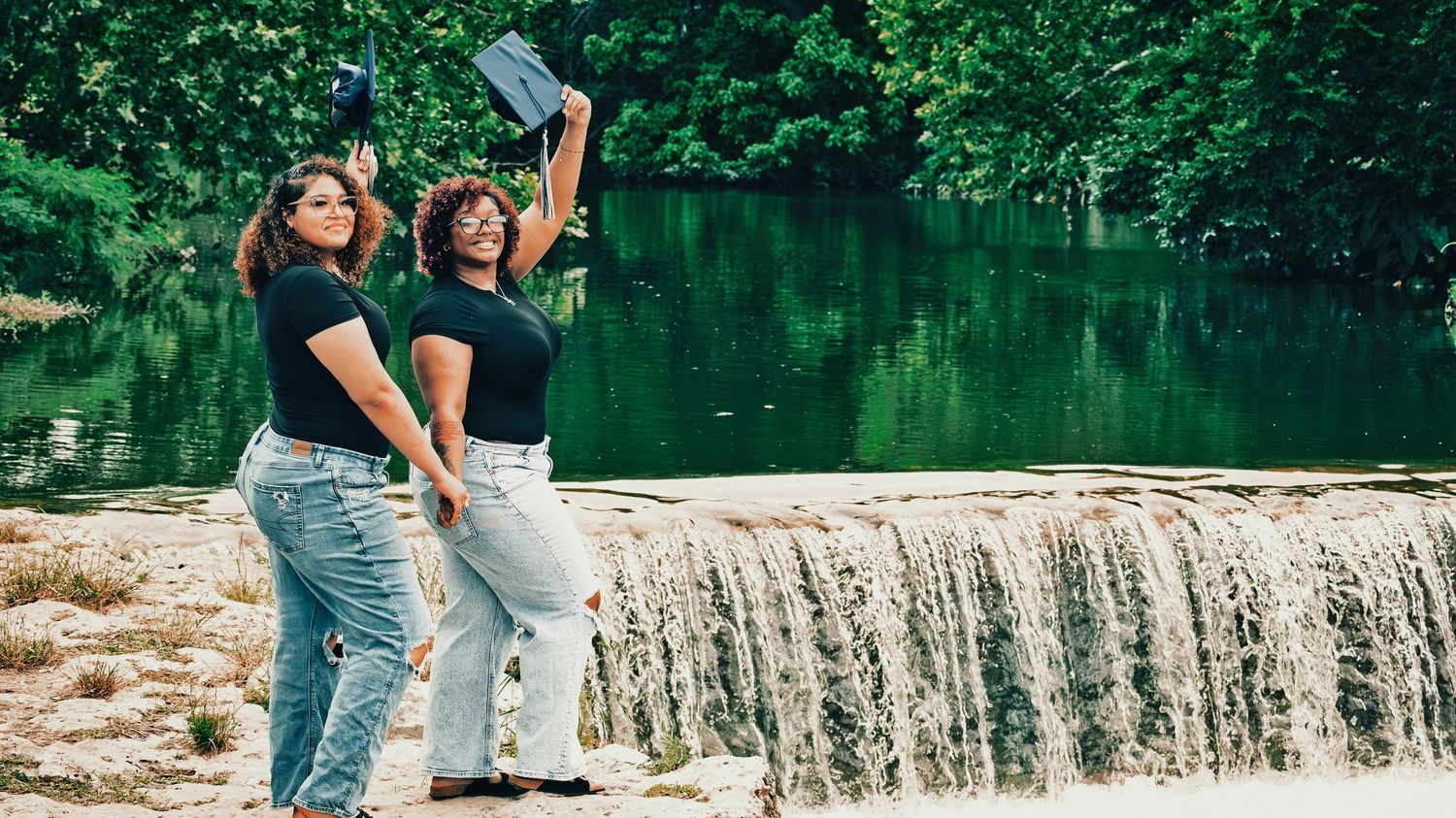 Two women in black tops hold graduation caps near a peaceful green waterfall backdrop for senior photos.