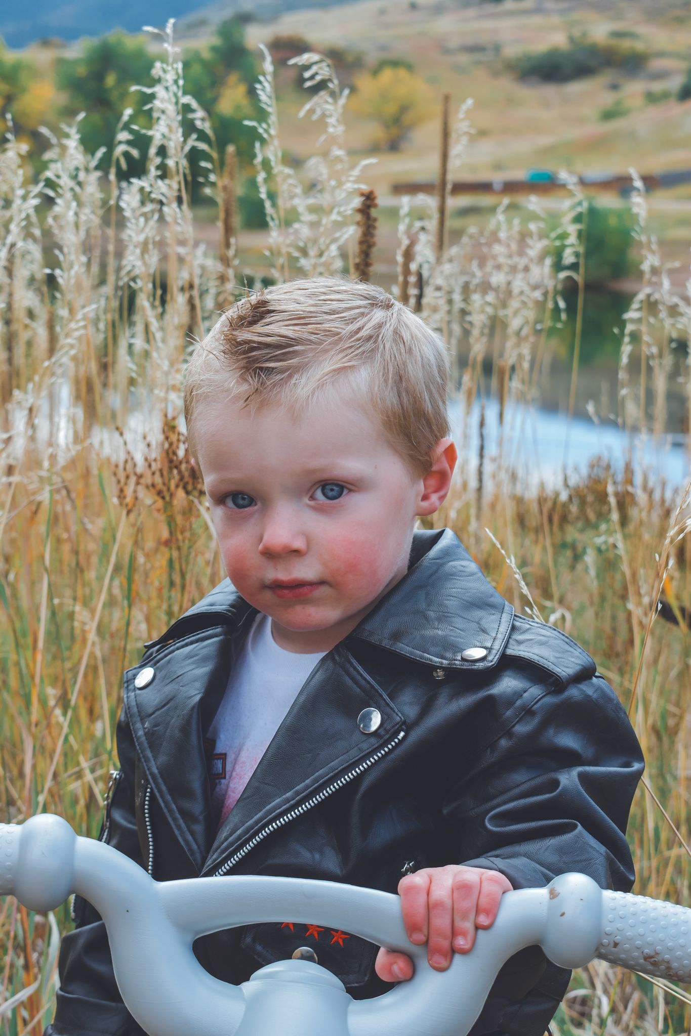 Serious blue-eyed toddler in black leather jacket standing by toy bike in tall grass field.
