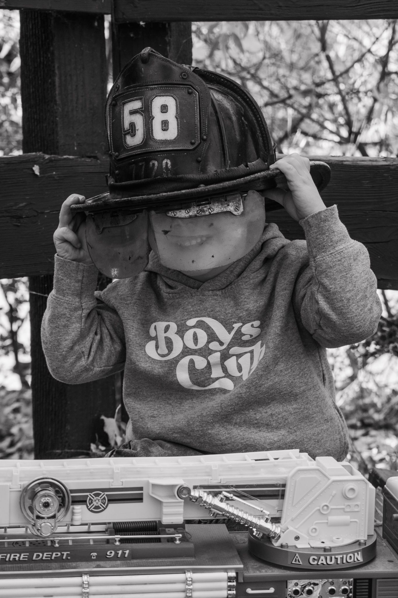 Black and white photo of boy in Boys Club sweatshirt wearing firefighter helmet number 58 with toy fire truck.