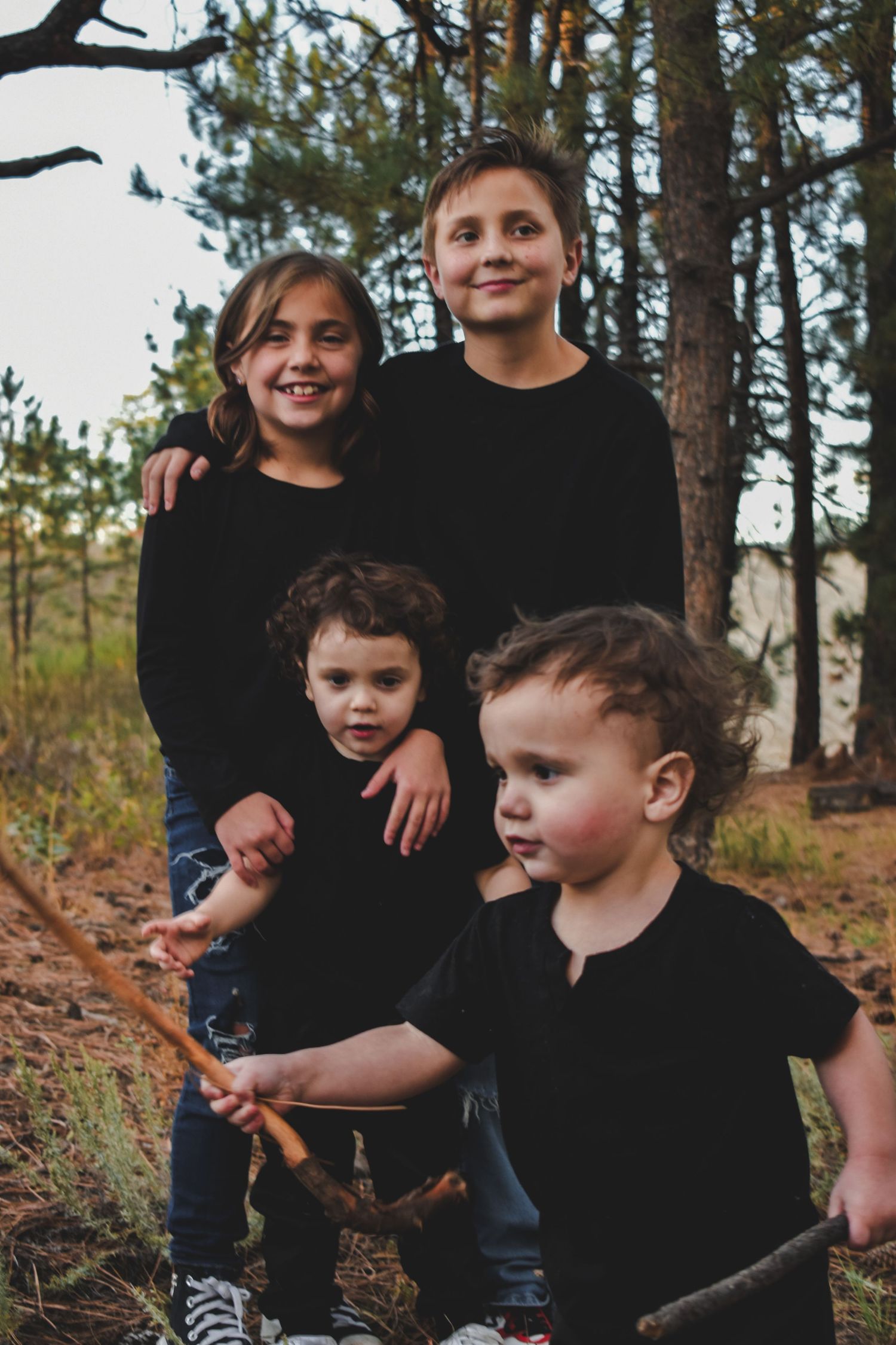 Four children in black outfits posing together among pine trees in a forest setting.