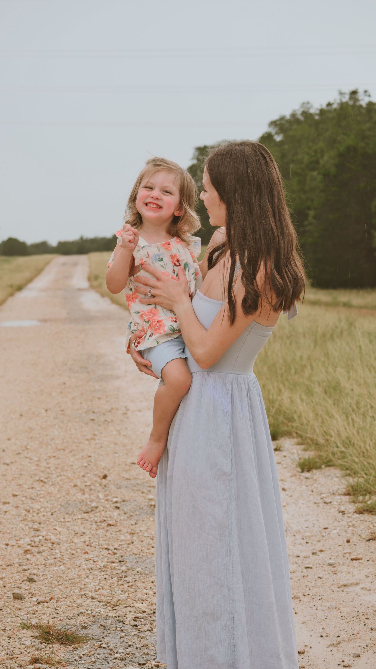 Smiling mother in gray maxi dress holds laughing little girl on a sunny dirt road.
