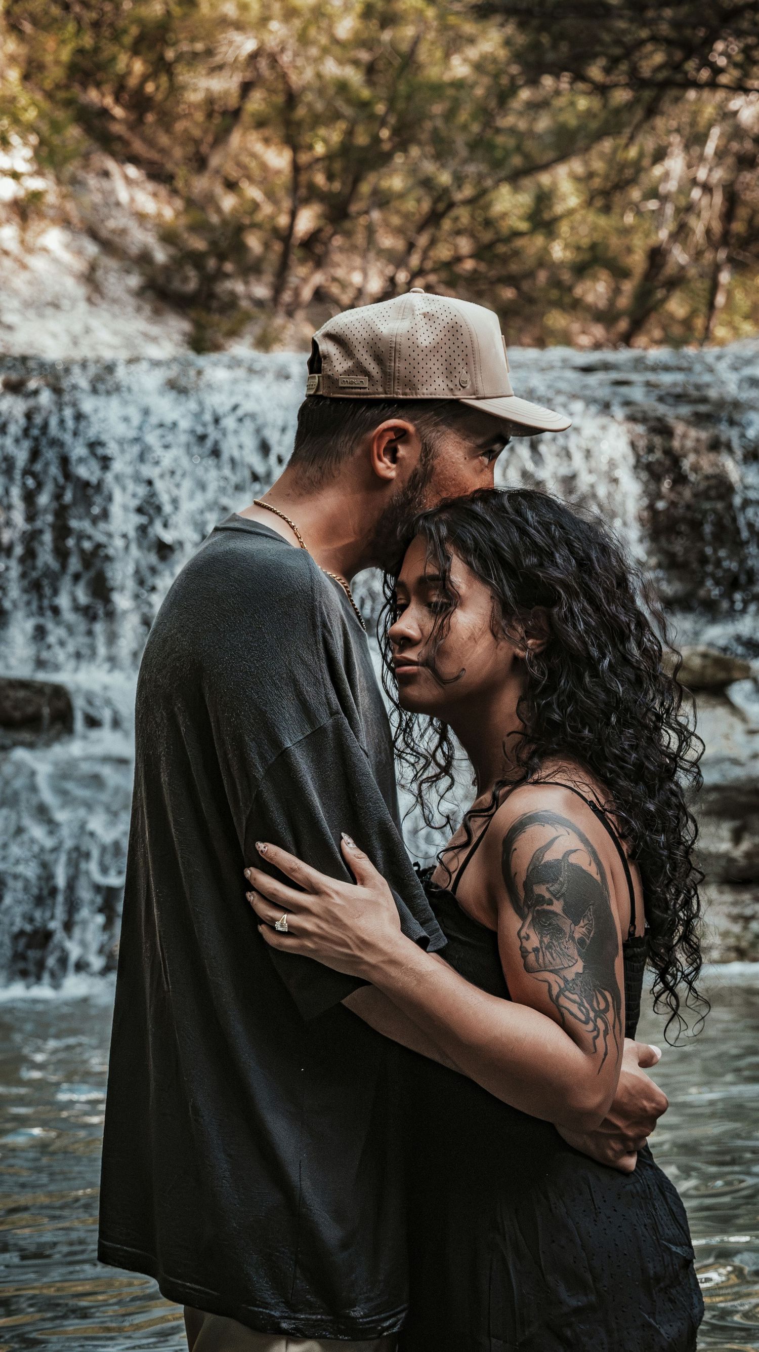 Tattooed couple hugging tenderly in front of a scenic waterfall.