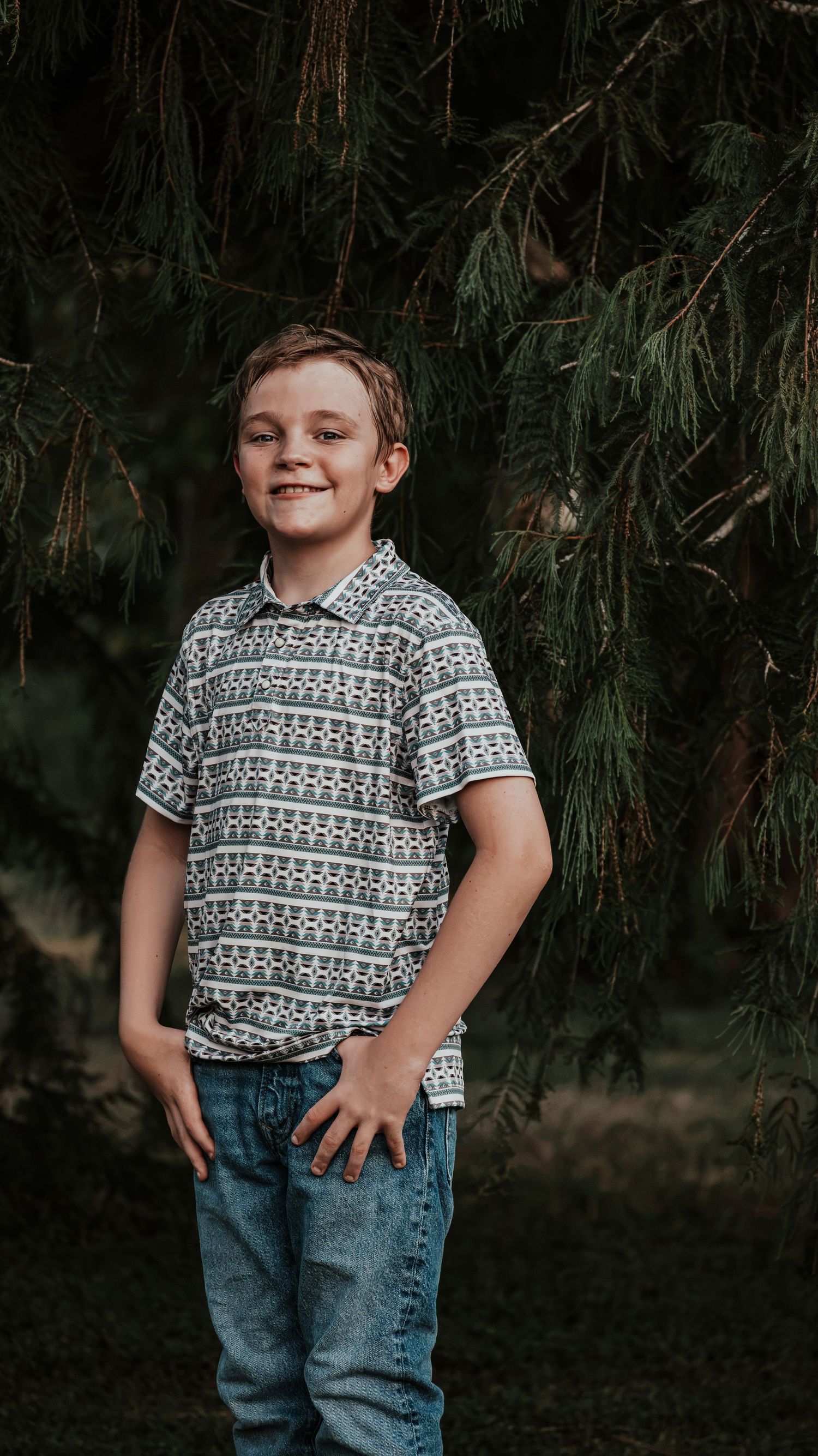 Smiling young boy in patterned polo shirt and jeans posing in front of evergreen trees outdoors.