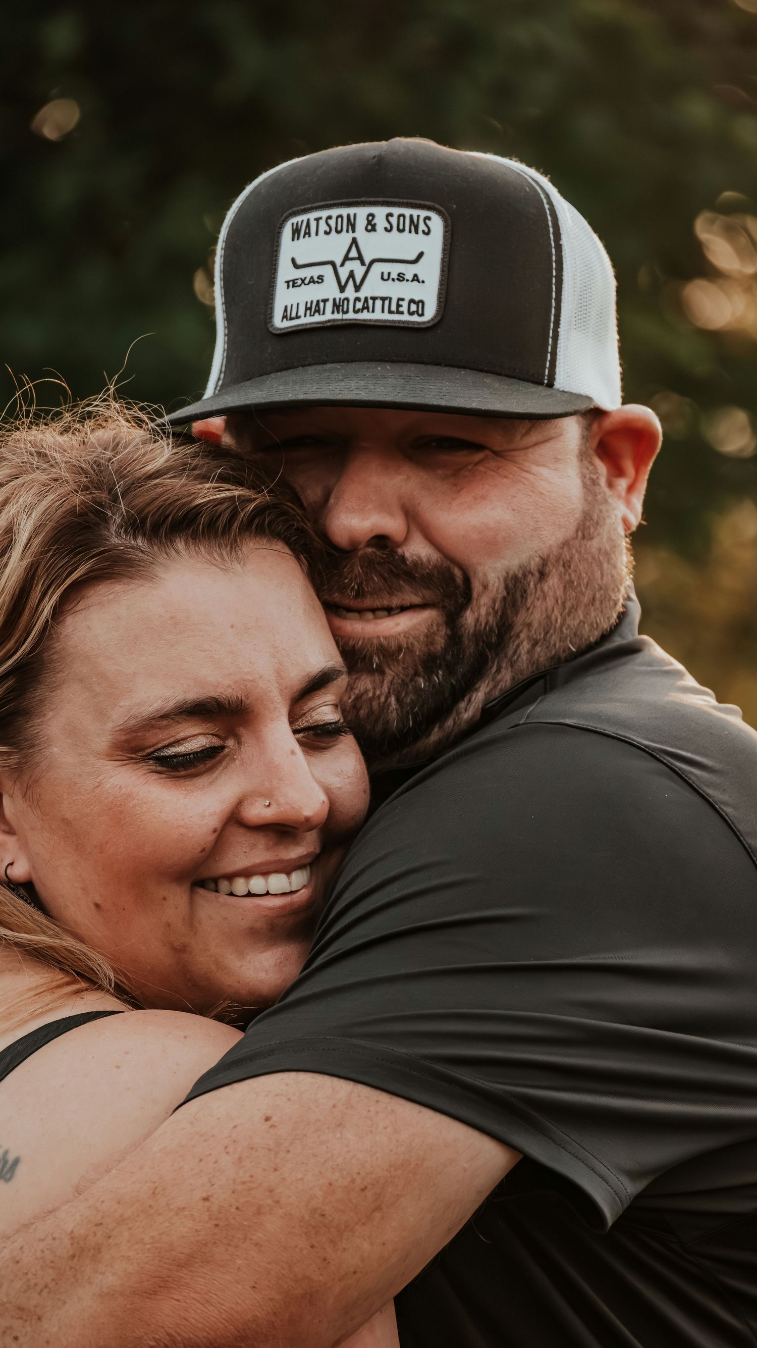 Couple sharing a warm hug outdoors, man wearing a Watson and Sons trucker hat.