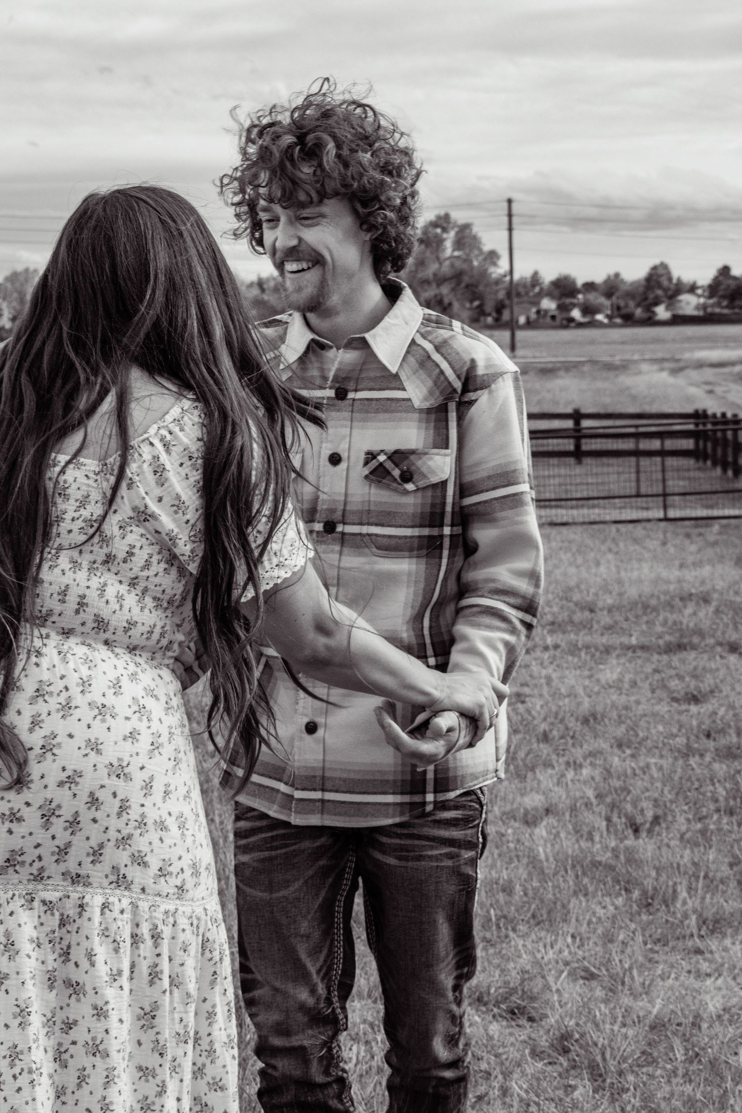 Black and white photo of a curly-haired man smiling at a woman in a floral dress on a farm.