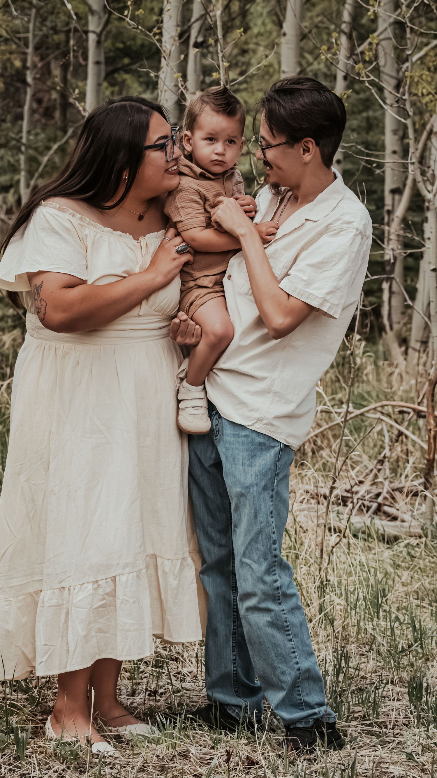 Loving parents in cream and white outfits hold their smiling baby in a peaceful wooded setting.
