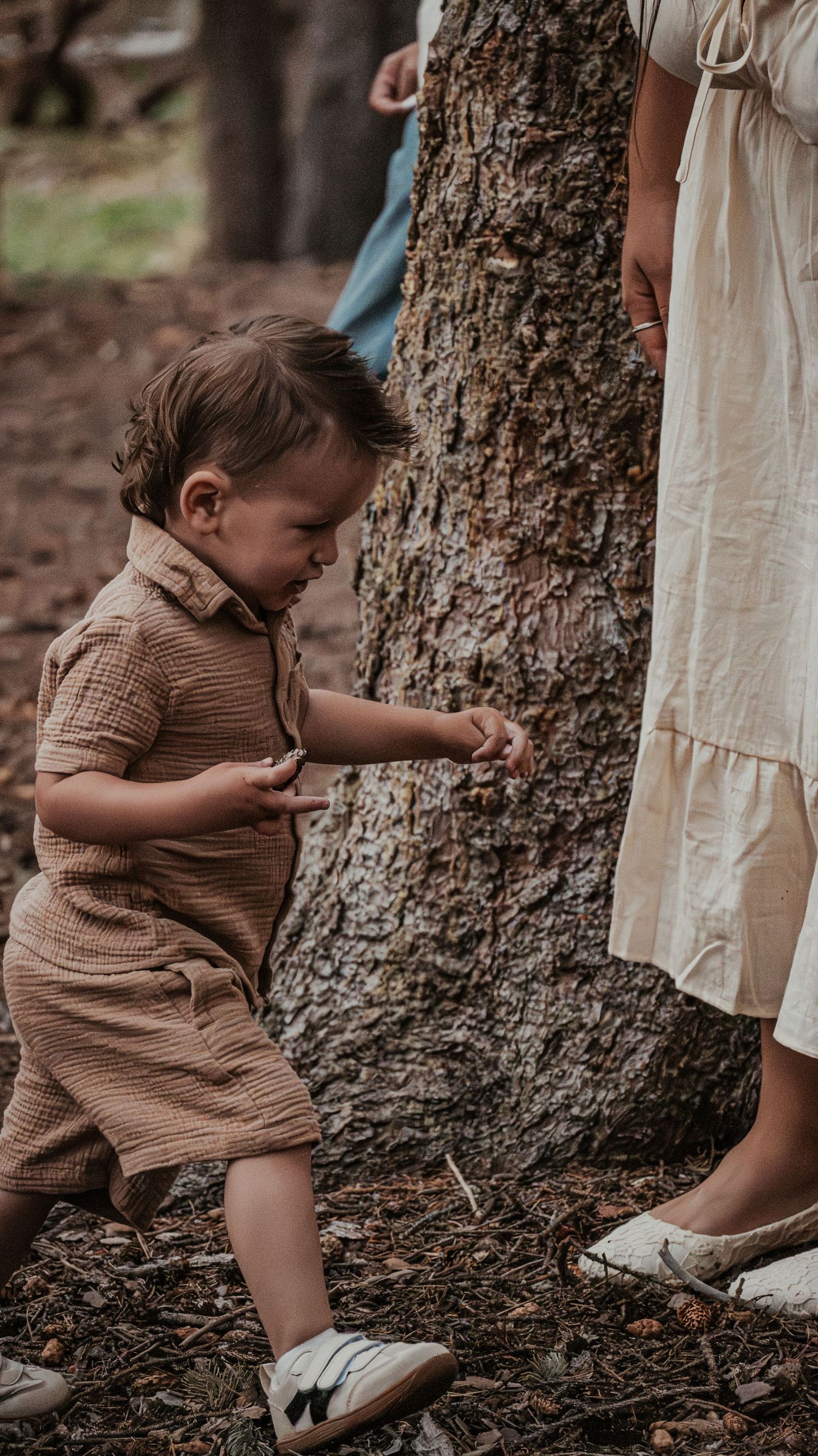 Curious toddler boy in brown linen set explores near a tree trunk beside his mother.
