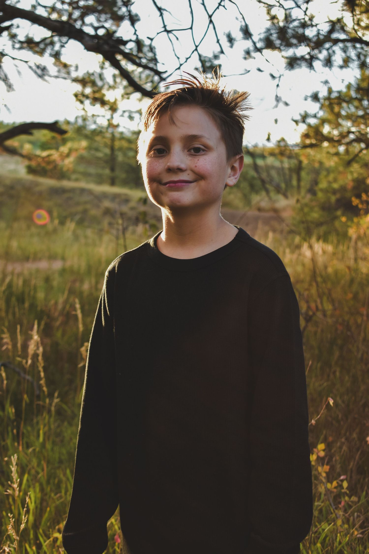 A smiling boy stands outdoors in a sunlit field with trees and green grass. Family Photographer in Pflugerville, Tx
