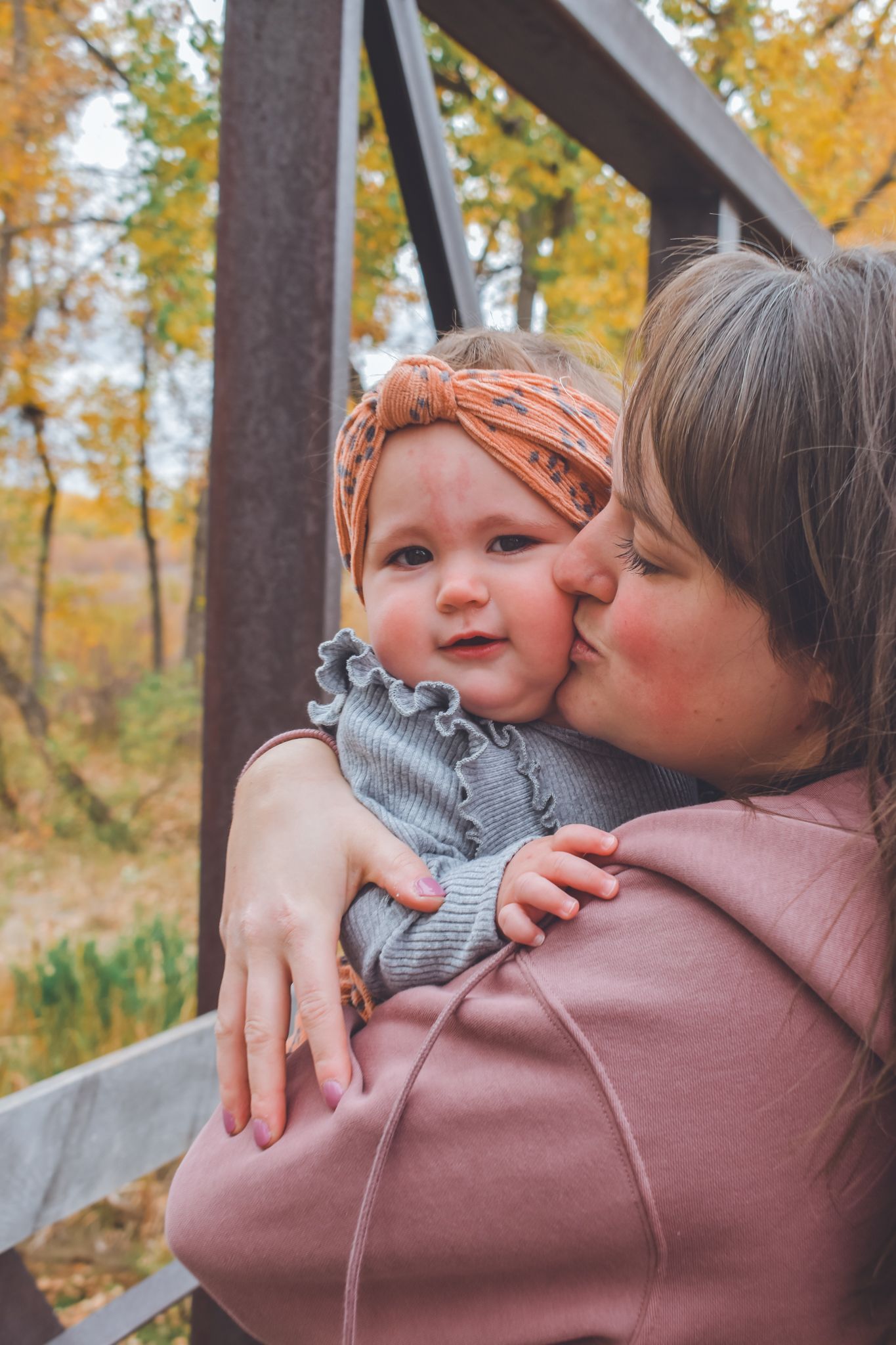 A mother tenderly kisses her baby girl wearing an orange headband outdoors in autumn. Family Photographer in Pflugerville, Tx