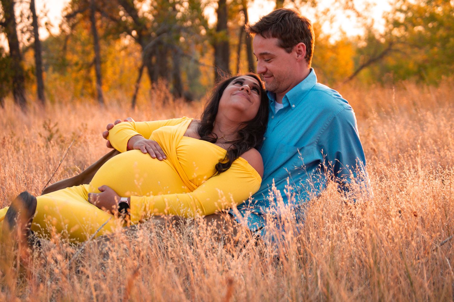 Expecting couple sitting together in tall golden grass field, woman in yellow dress leaning back against partner.