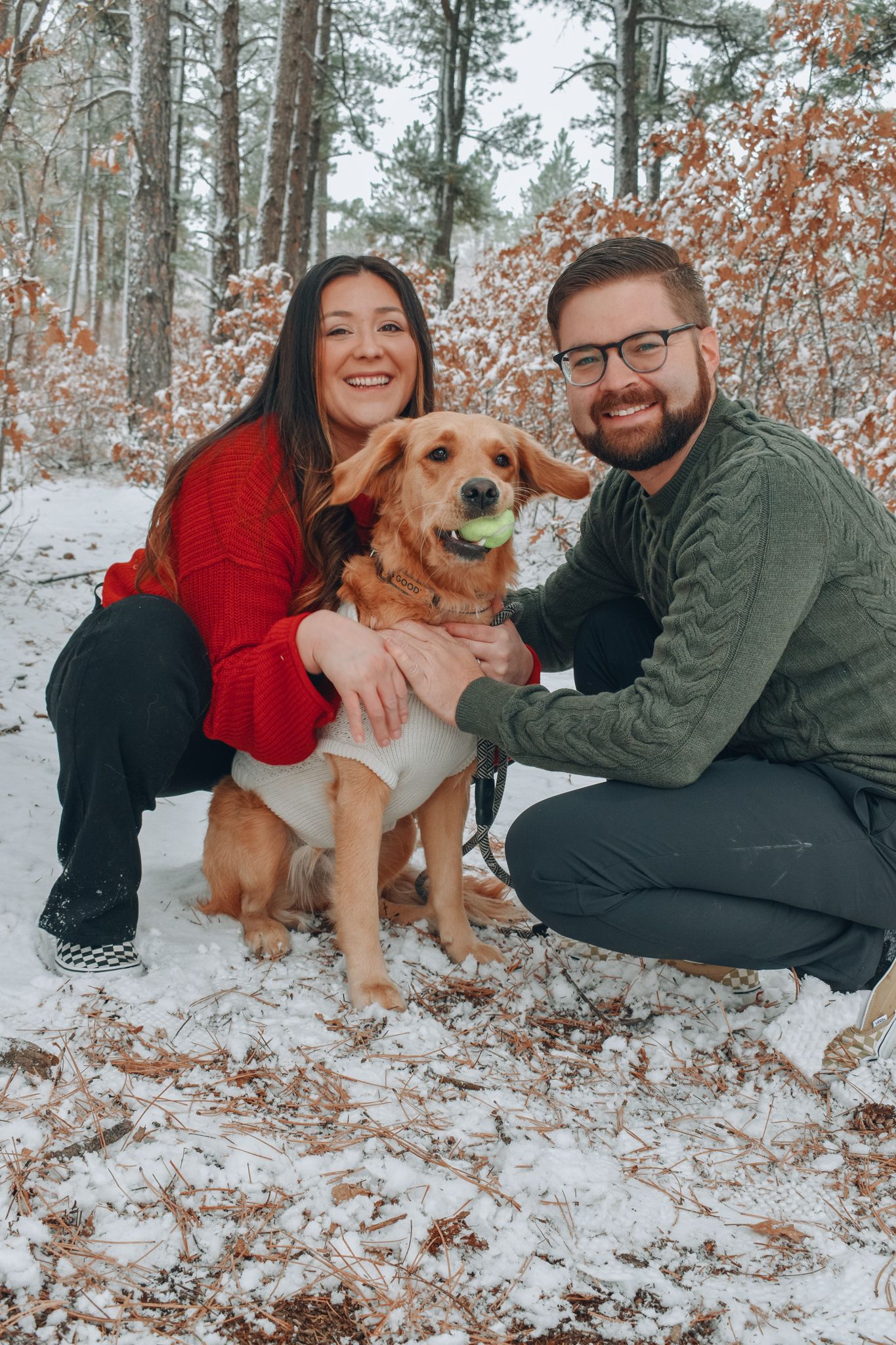 Smiling couple crouching beside their golden dog holding a tennis ball in a snowy forest.