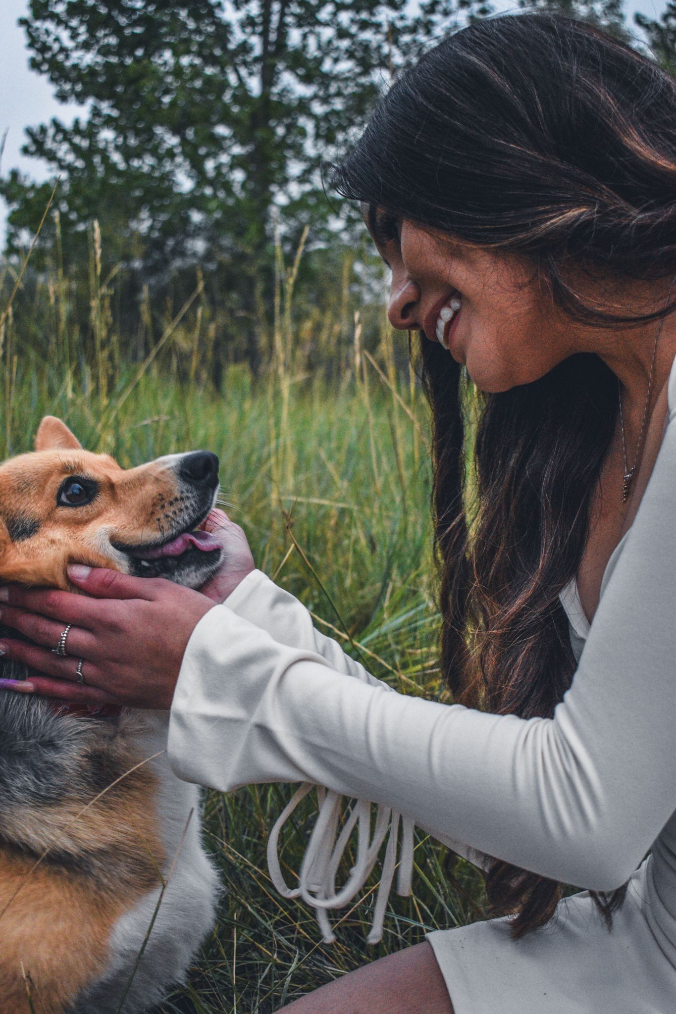 Smiling young woman with long dark hair cuddles a happy Corgi dog in a grassy outdoor field for senior photos.