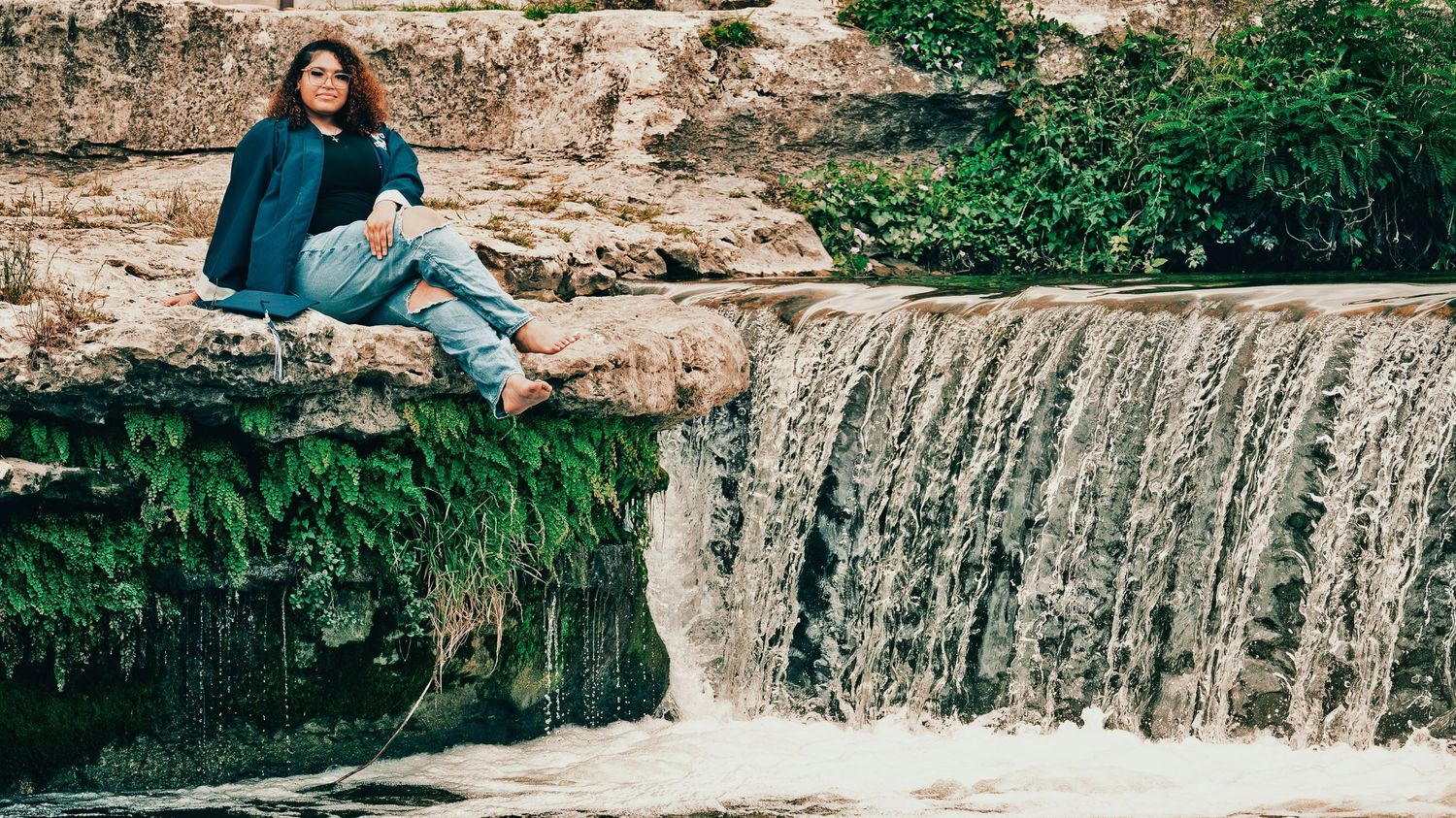 Young woman in blue jacket sitting on rocky ledge beside a small waterfall outdoors for senior photos.
