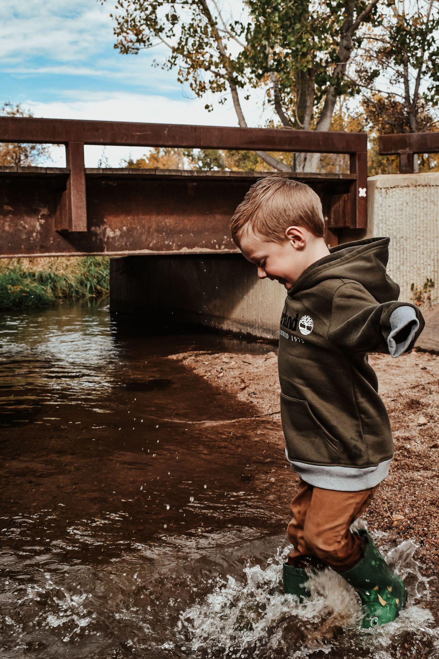 Young boy in green rain boots splashing in a shallow creek wearing a Timberland hoodie outdoors.