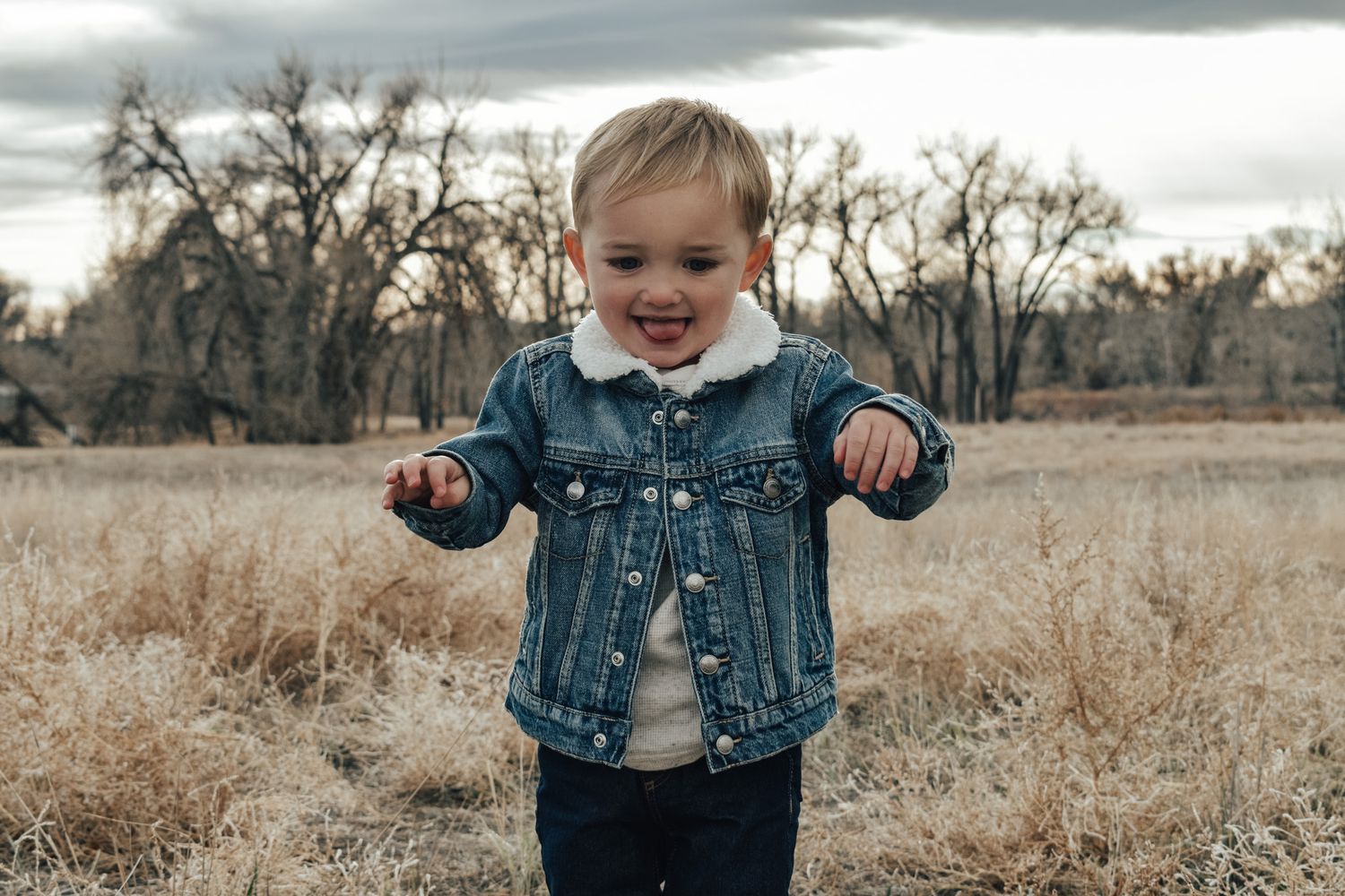 Happy toddler boy smiling in a denim sherpa jacket standing in a dry autumn field.