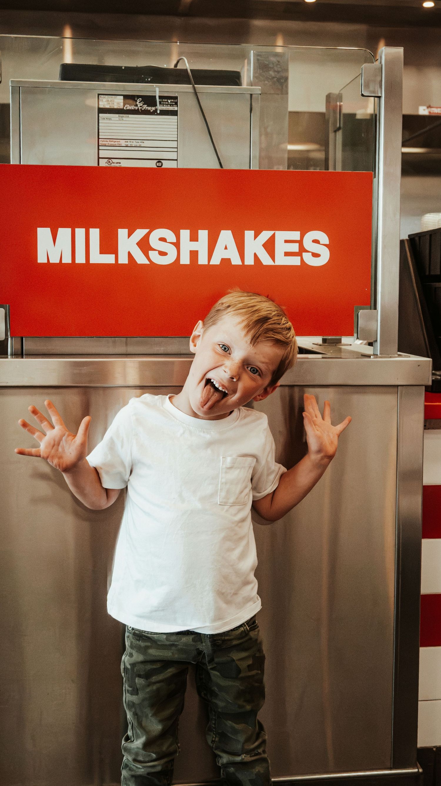 Excited young boy in white tee and camo pants sticking out tongue in front of a red Milkshakes sign. Family Photographer in Pflugerville, Tx