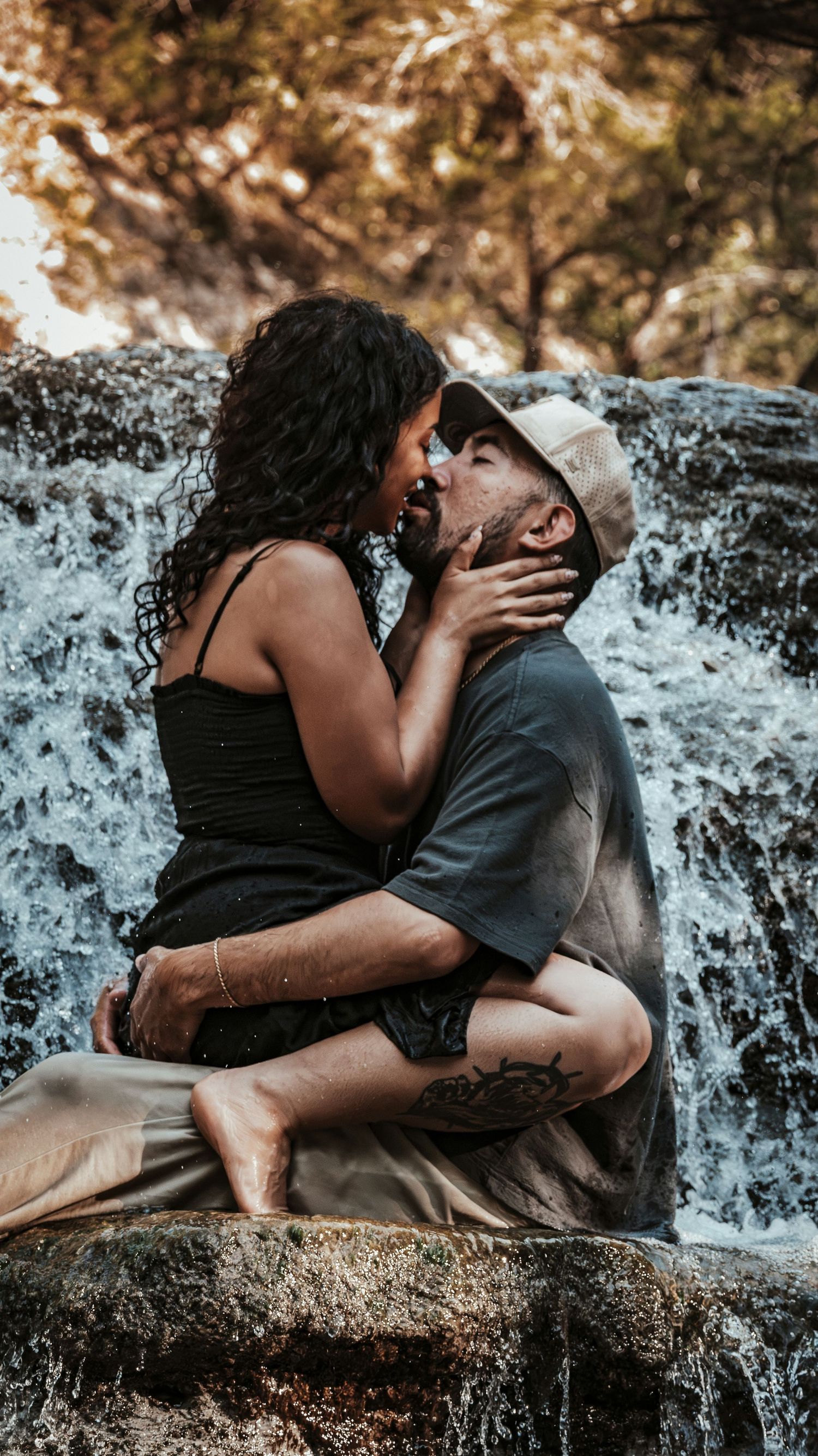 Romantic couple sharing a kiss while sitting on rocks in front of a waterfall for a couples session.