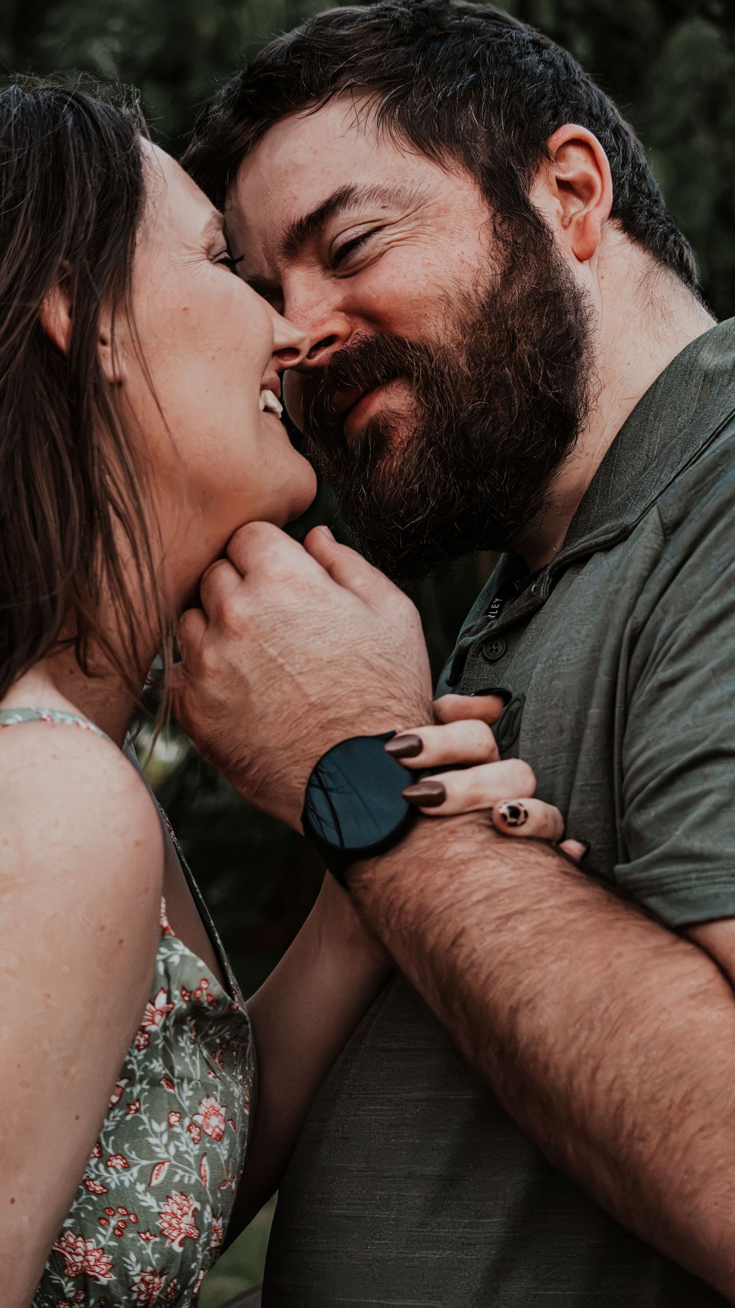 Couple sharing an intimate moment, smiling and almost kissing outdoors, man with beard wearing a watch, woman in floral dress.