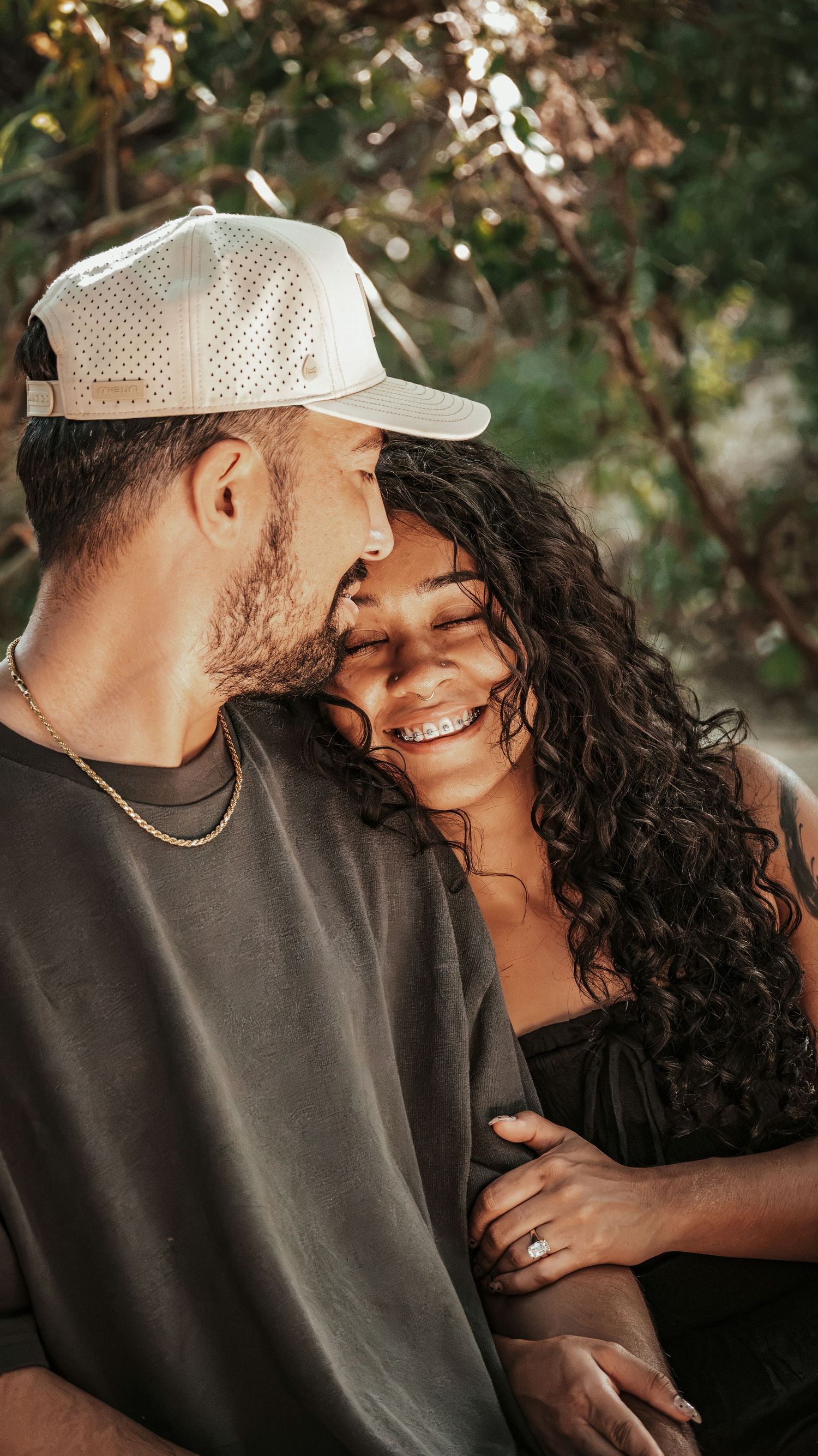 Man in white snapback cap nuzzling smiling woman with curly hair and tattoo outdoors in warm sunlight.