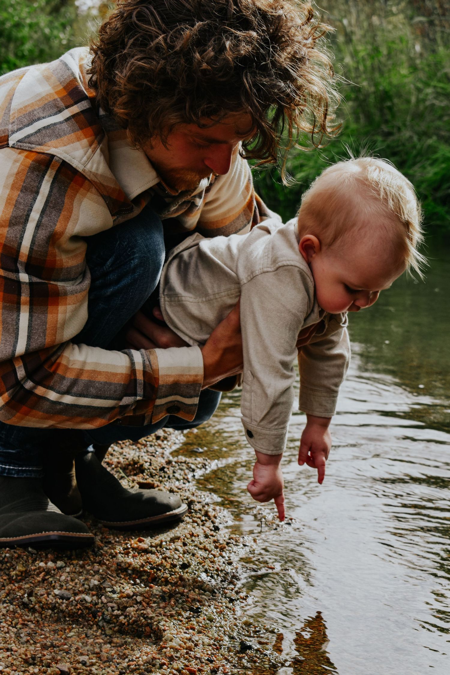 Father in plaid shirt helping curious blonde toddler touch water at river's edge outdoors.