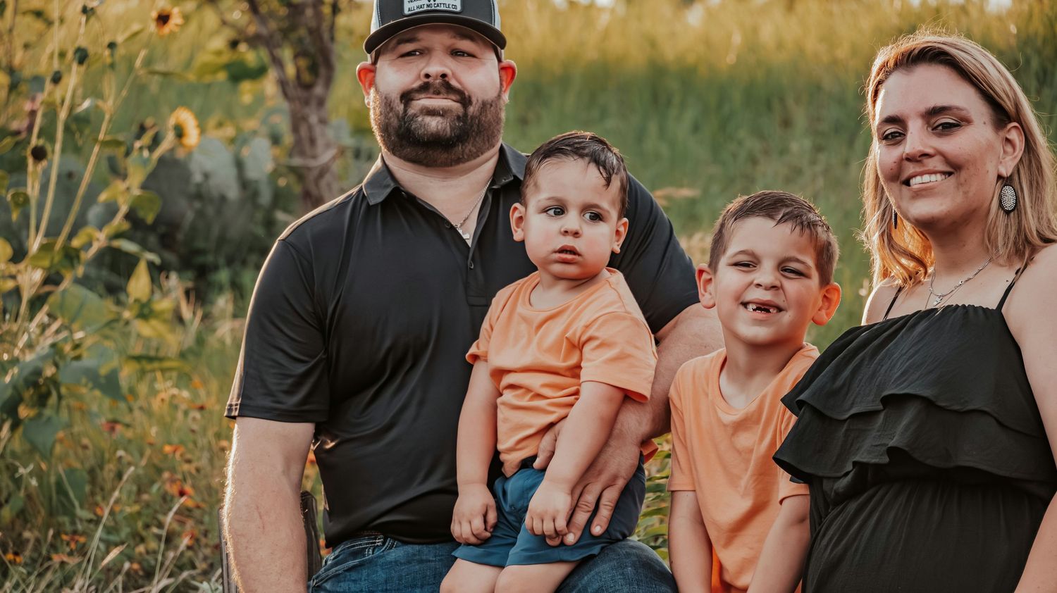 Family of four sits together outdoors, dad holding toddler in orange shirt while mom and older son smile.